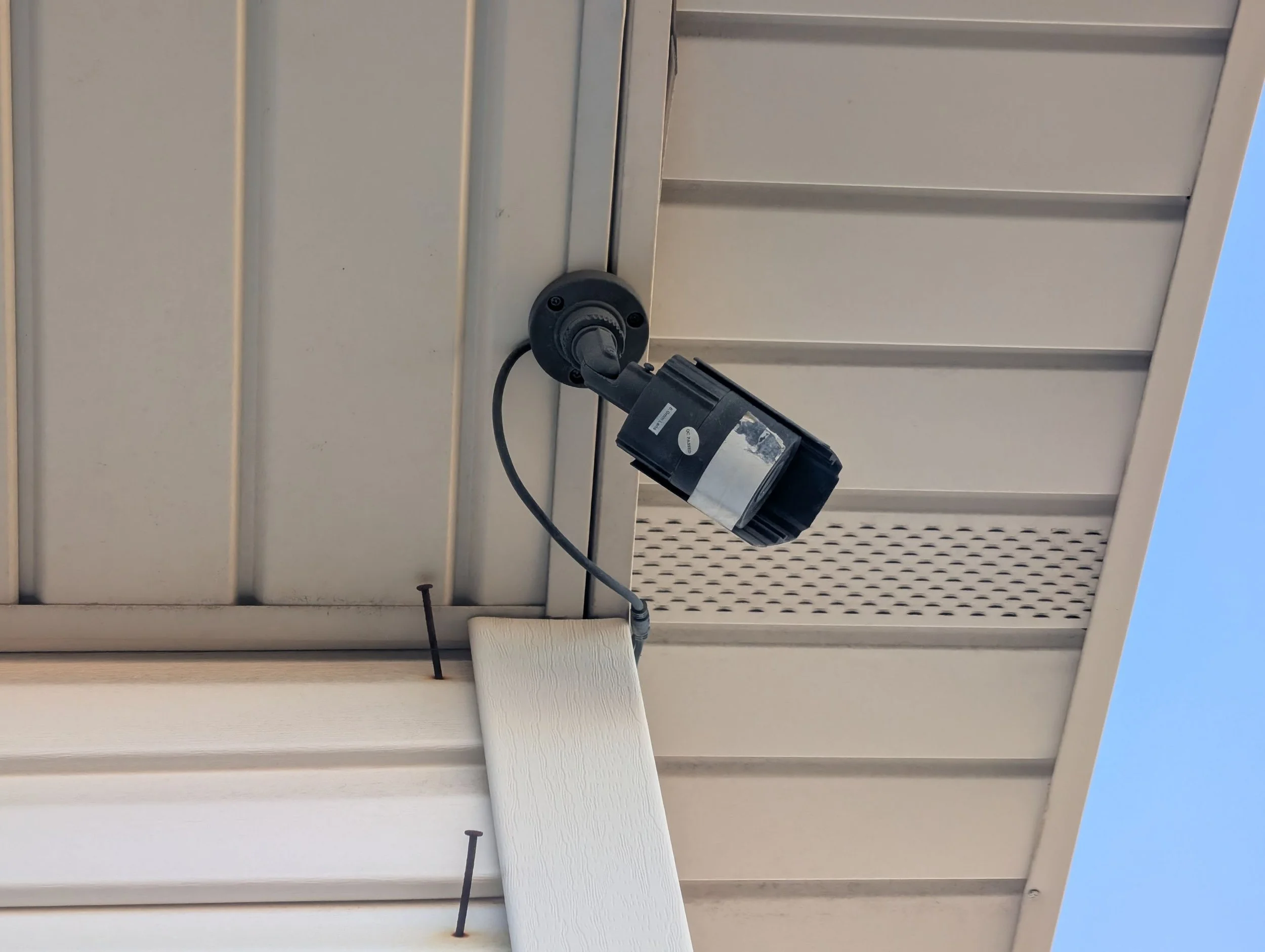 Exterior of a building showing a wall-mounted security camera pointing downward, attached to a white tiled wall with a blue sky visible at the edge.
