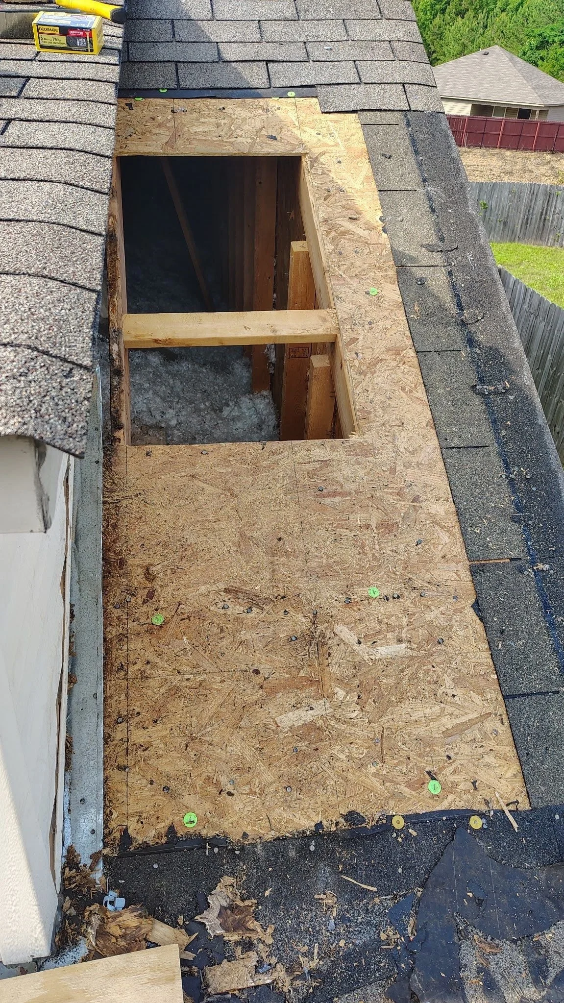 Roof with an opening cut out for a skylight, with plywood and framing installed around the opening, on a house with shingles, on a sunny day, with neighboring houses and greenery in the background.
