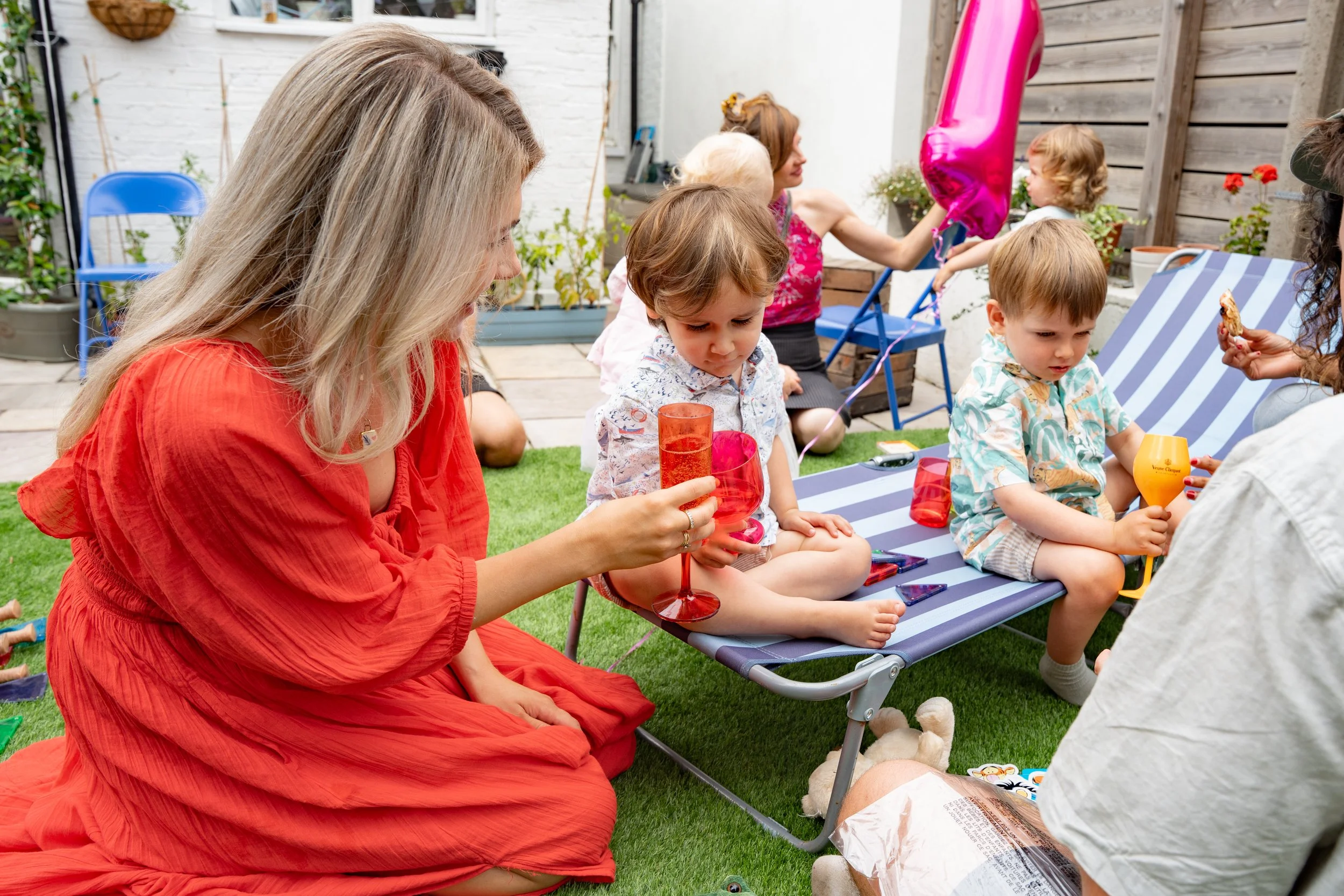 People celebrating a child's birthday in a backyard. A woman in a red dress is sitting on the grass, holding a drink. Two children are sitting on a striped outdoor bench, one with a pink balloon and the other with a yellow toy. Other women and childr