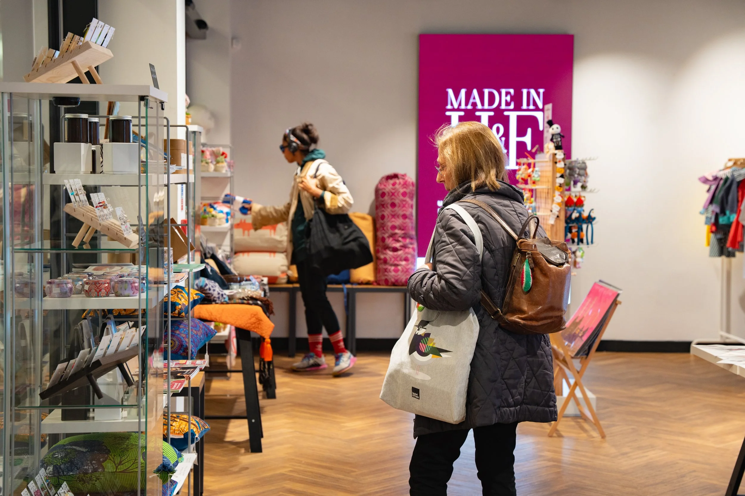Two women shopping in a store with various home decor and gift items on display, including pillows, candles, and small figurines, with a pink sign reading 'Made in T&F' in the background.