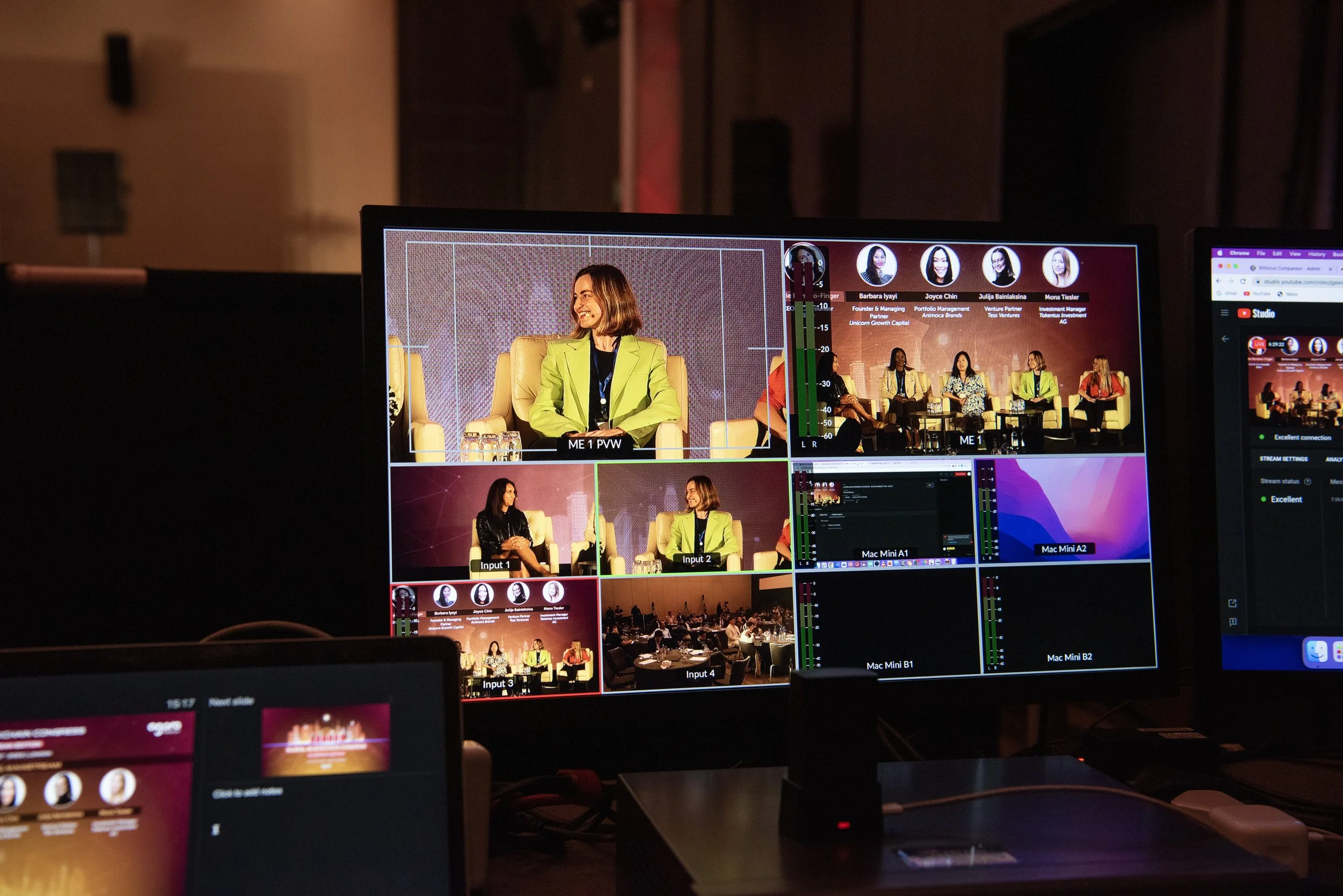 A video control room with multiple monitors showing a panel discussion with several women speaking on stage.
