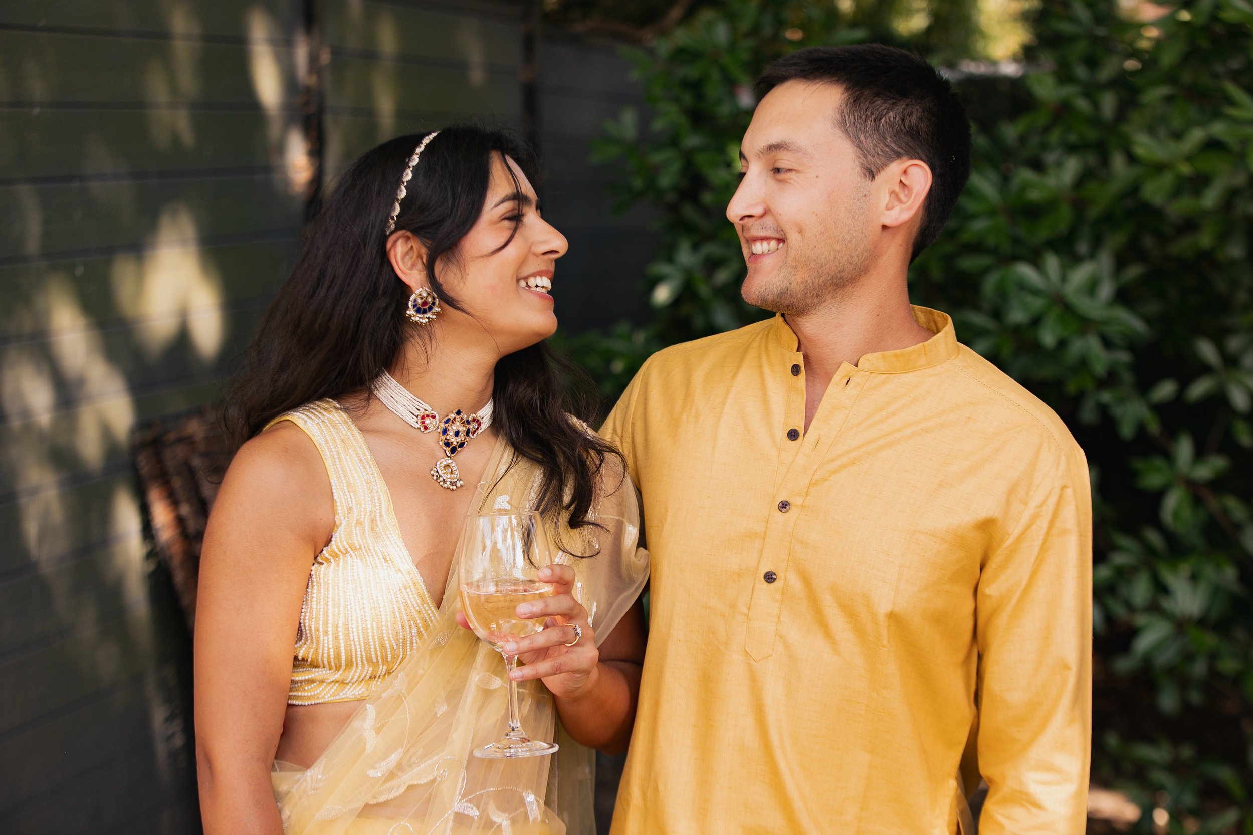 A woman in a yellow dress holding a wine glass, smiling and talking with a man in a yellow shirt outdoors with green foliage in the background.
