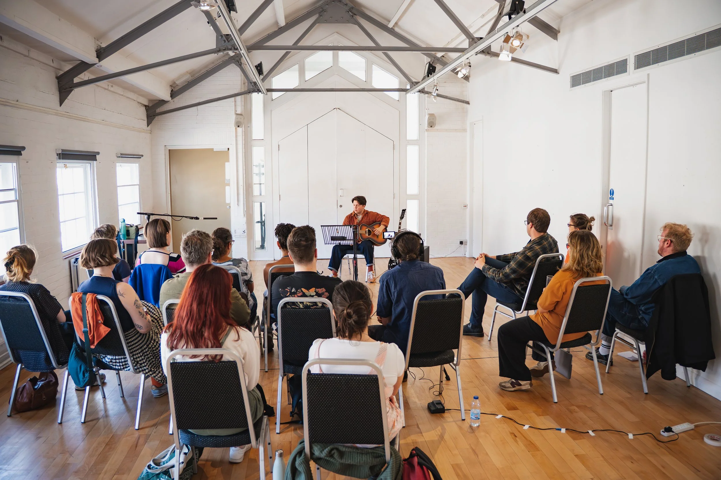 A person playing guitar and singing in front of seated audience in a bright, spacious room.
