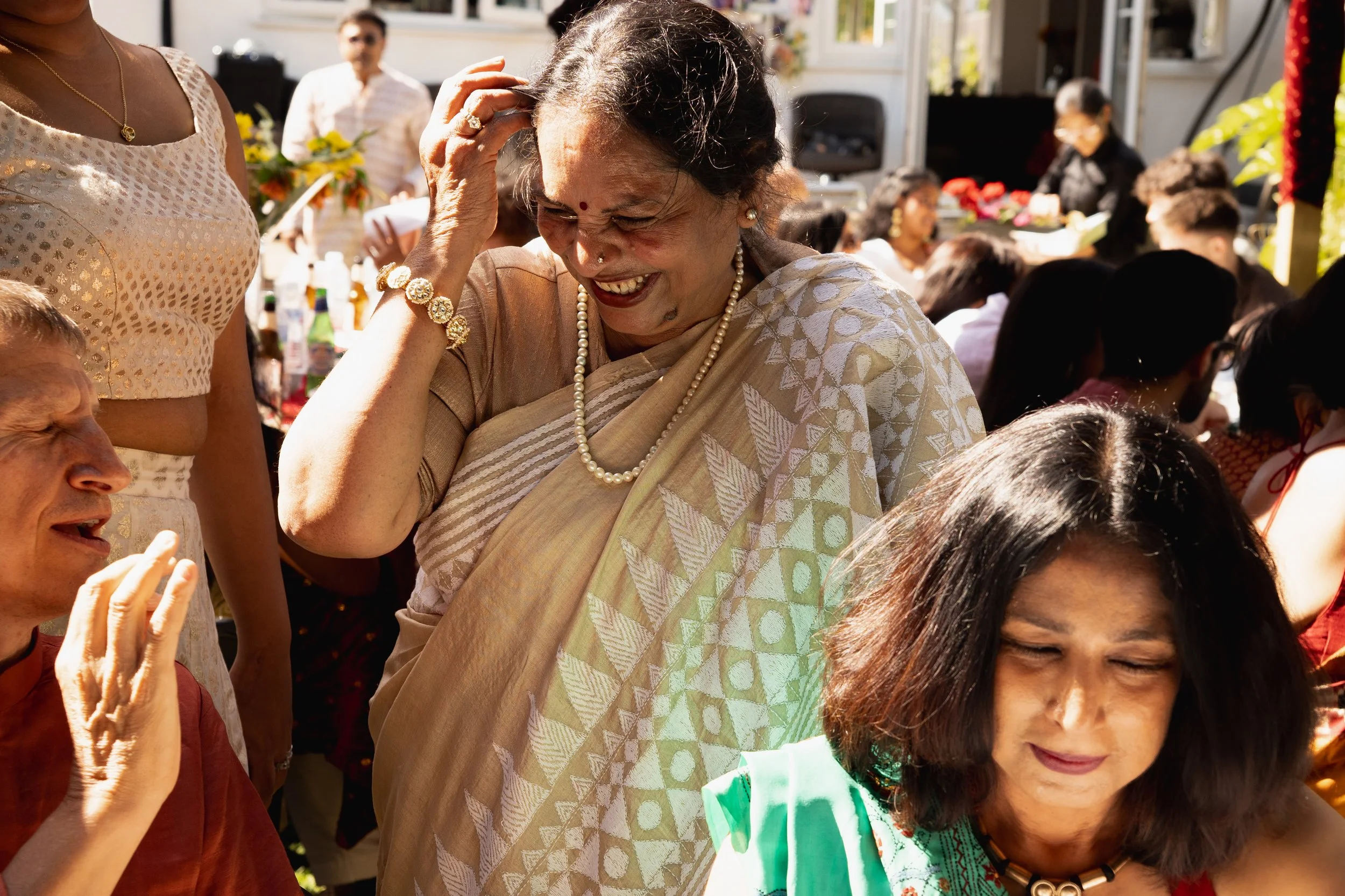 A woman in traditional Indian attire laughing and smiling at a lively social gathering, surrounded by other people in festive clothing.