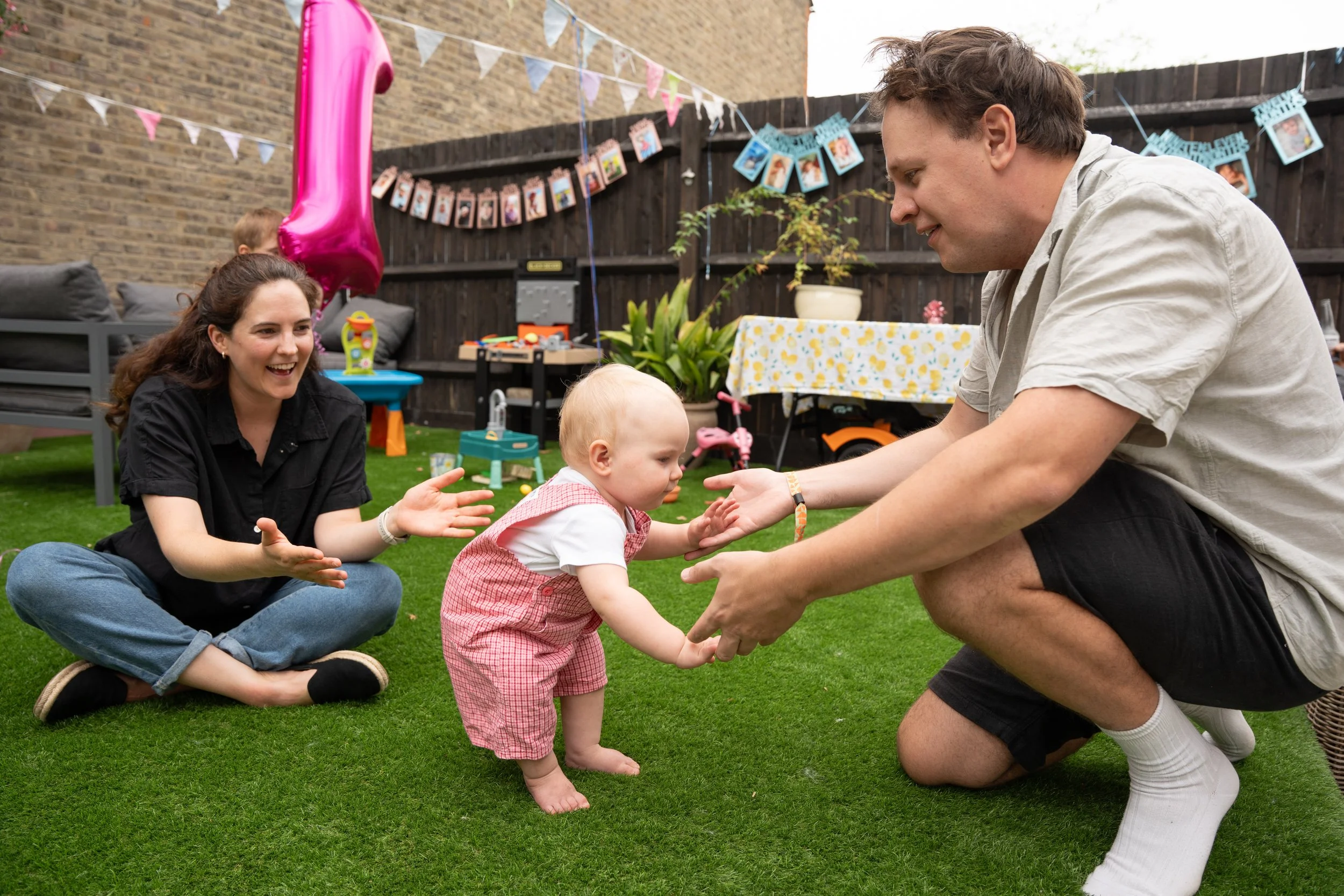 A family celebrating a child's birthday in a backyard. The child, in pink checkered overalls, is standing and holding hands with a man who is crouching. A woman is sitting on the grass, smiling and clapping. The backyard is decorated with balloons, b