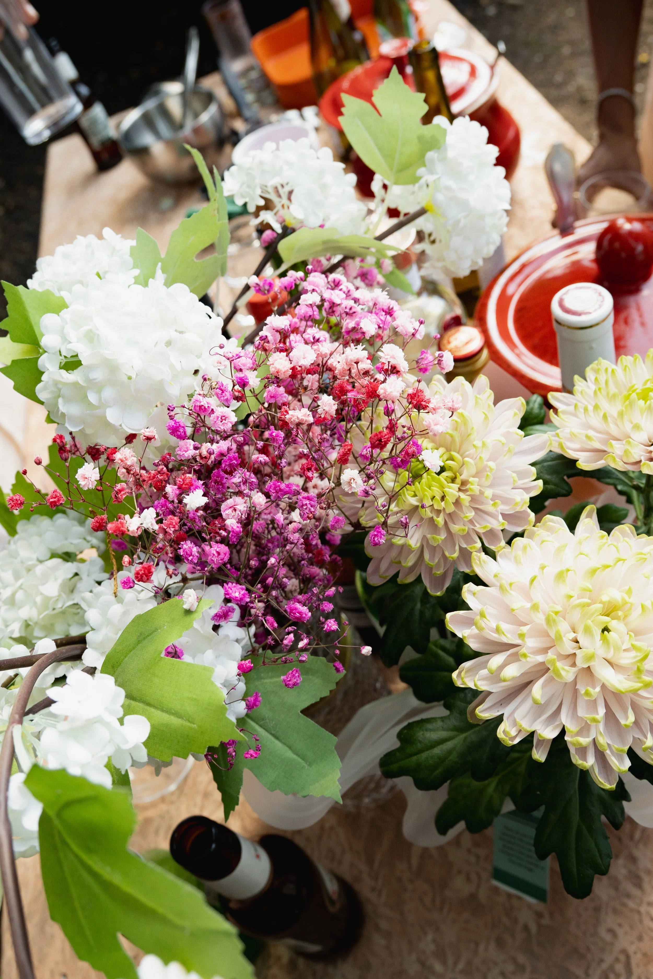 A collection of white, pink, and cream-colored flowers in vases on a table, with various bottles and kitchen items in the background.