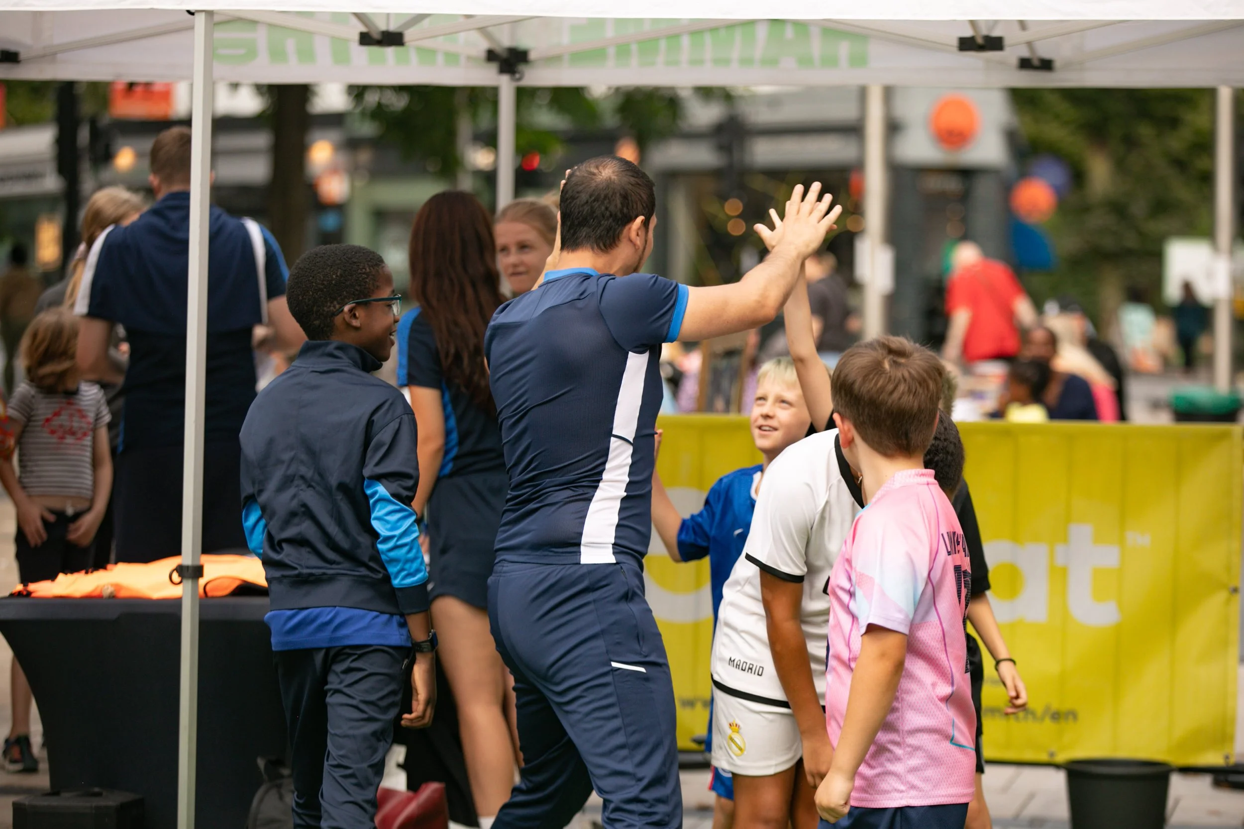 A group of kids and a man in a blue sports shirt high-fiving under a white canopy at an outdoor event. Kids are smiling, and there are booths and other people in the background.