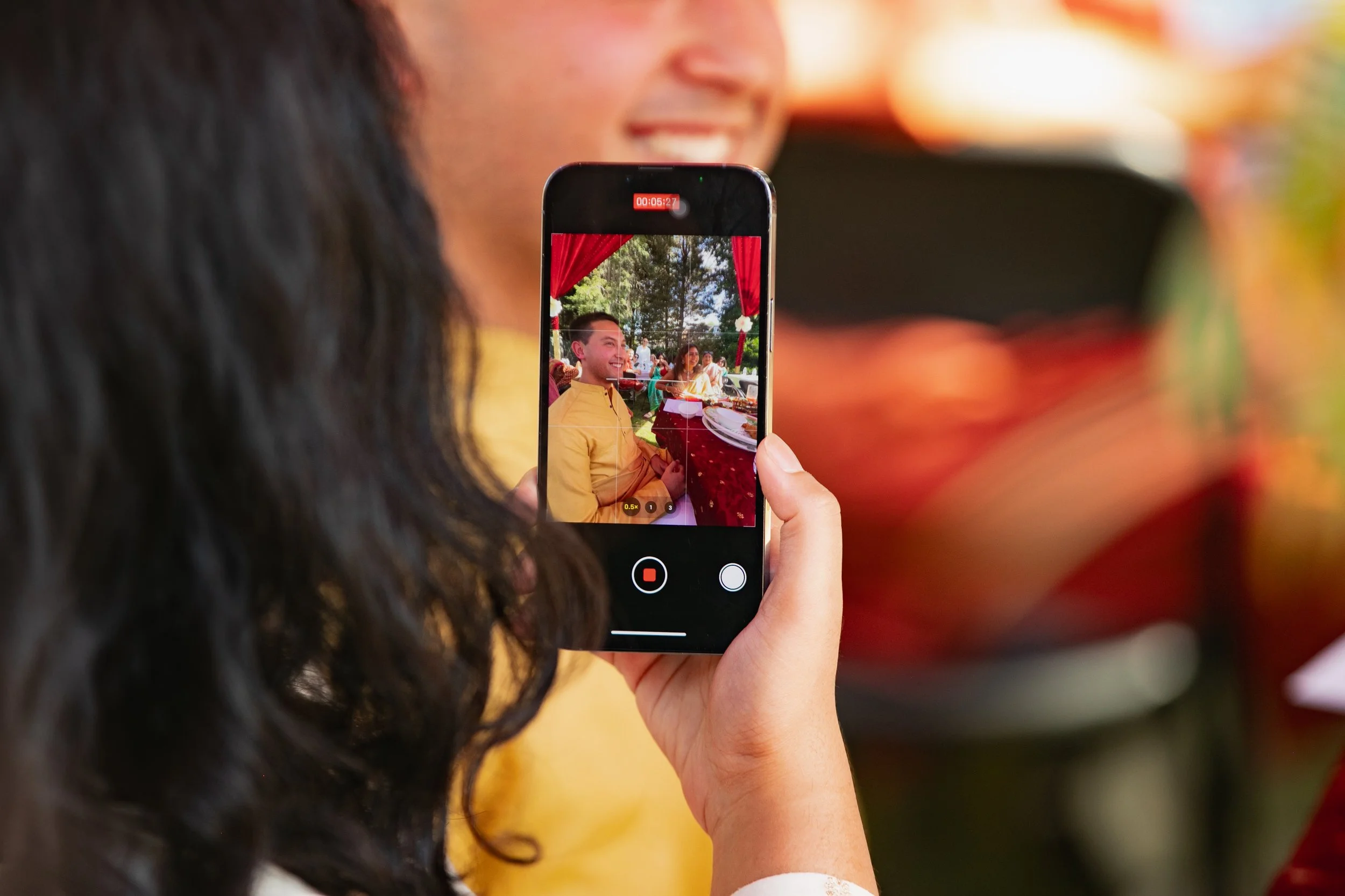 Person taking a photo or recording a video of a smiling man in a yellow shirt at an outdoor event with red curtains and trees in the background.