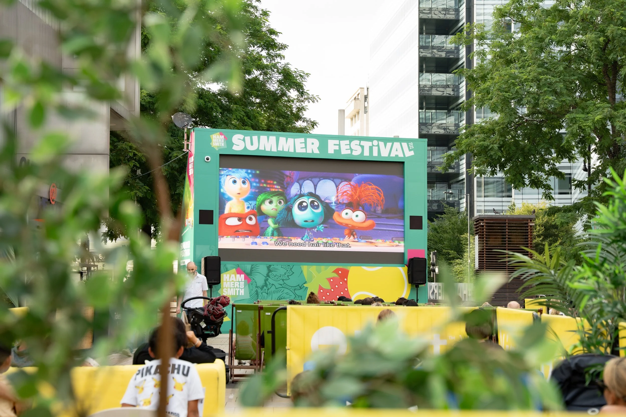A large outdoor screen at a summer festival showing animated characters from the movie 'Inside Out' with a subtitle that says, 'We need hair like that.' The screen is framed by a green border with "SUMMER FESTIVAL" at the top and surrounded by trees 