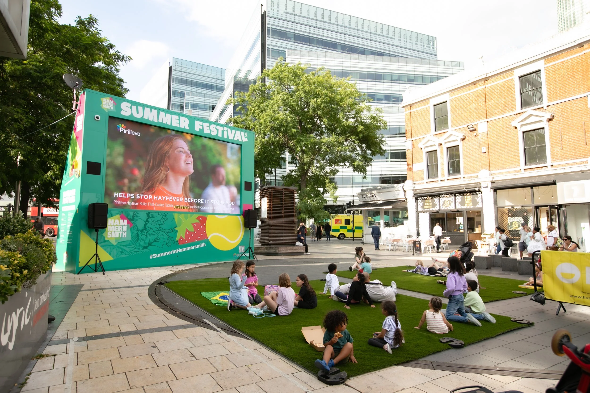 People sitting on artificial grass in an outdoor urban plaza watching a large outdoor screen displaying a woman smiling, promoting a summer festival in Hammer Smith.