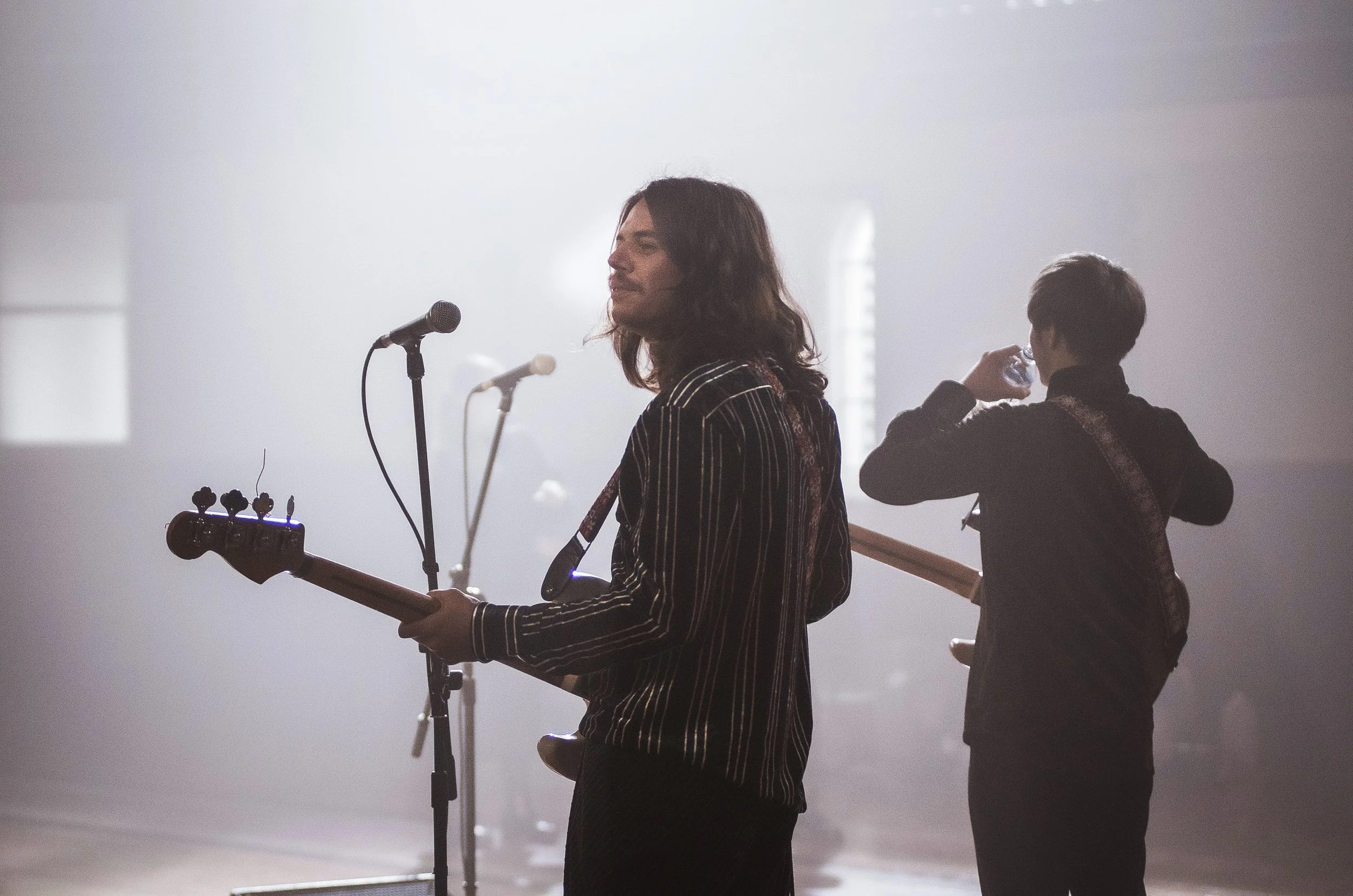 Two musicians on stage, one playing a guitar and the other drinking water, with microphones and stage lights in the background.