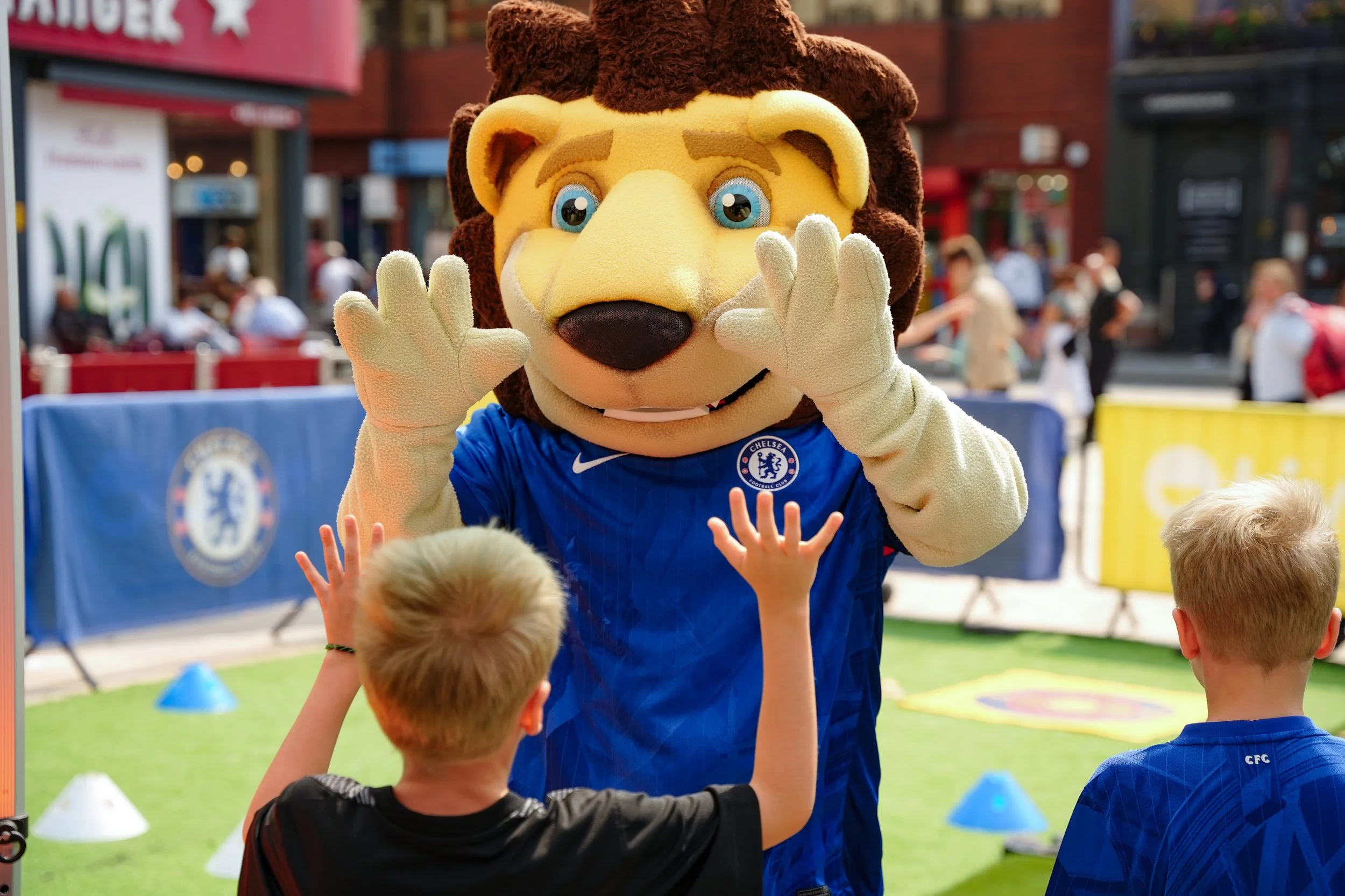 A person dressed as a lion mascot wearing a Chelsea Football Club jersey, high-fives two young boys at an outdoor event.