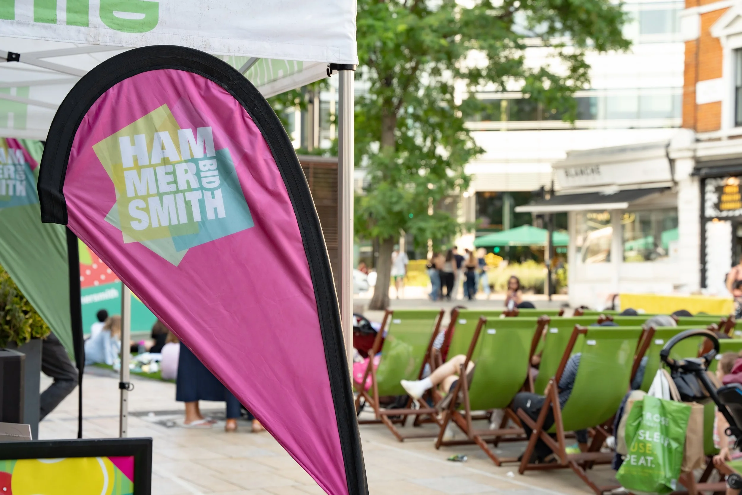 A pink flag with the text for the Hamber Smith Big logo on it, set up at an outdoor event with people sitting on green chairs and walking in the background.