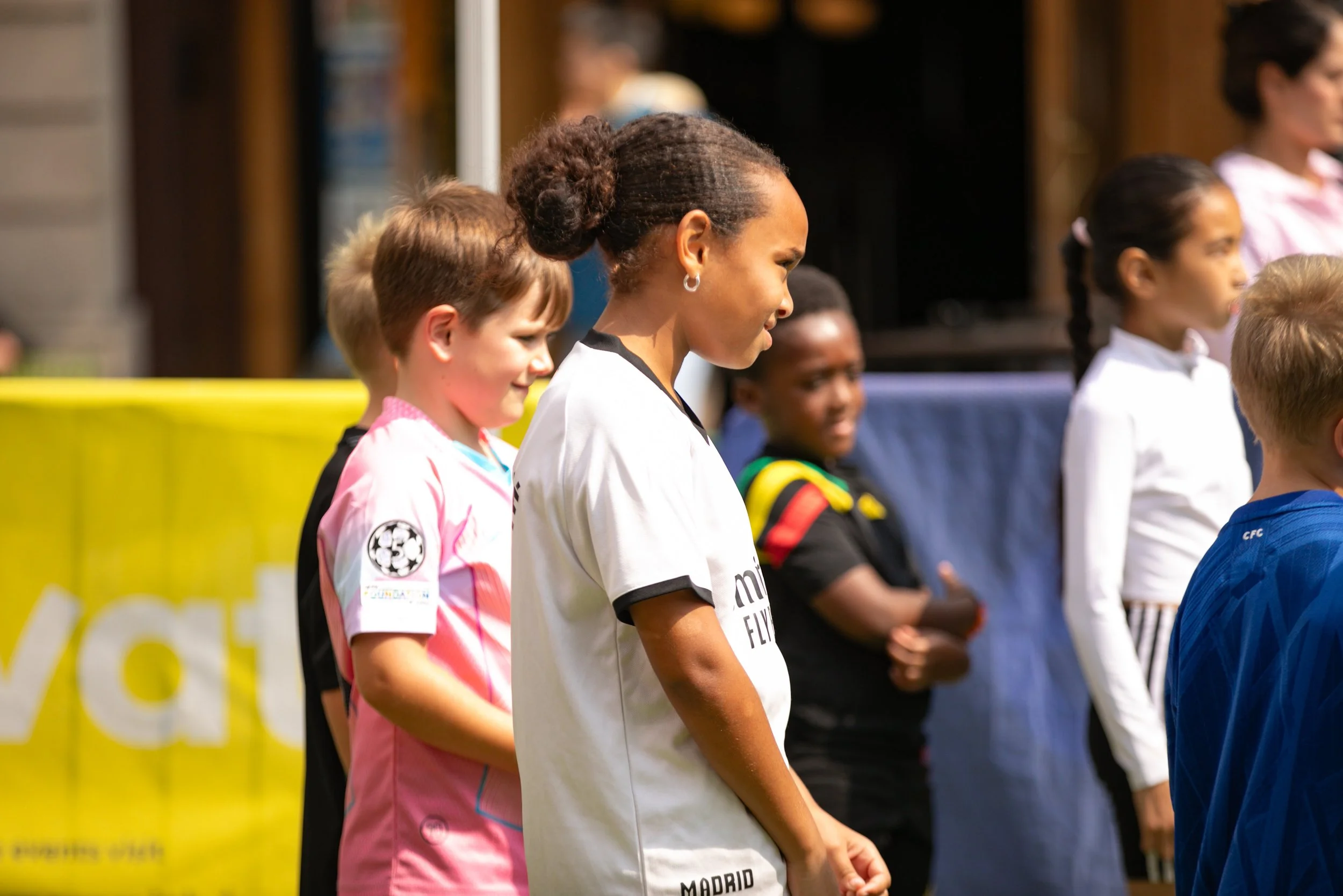 Children standing outdoors, some wearing sports jerseys, in a line, with a yellow fence and a building in the background.