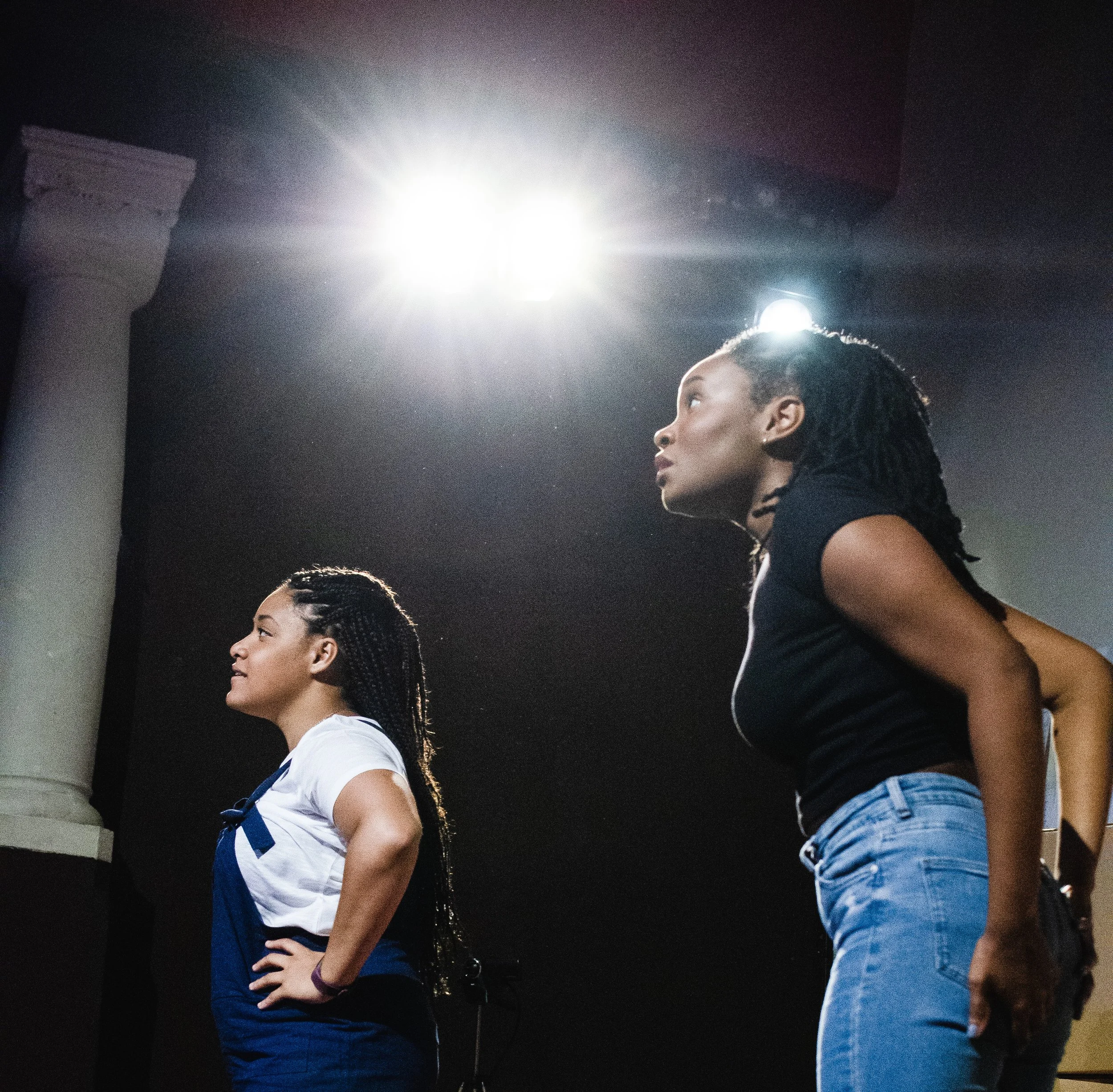 Two young women standing side by side, facing the same direction, with a bright light overhead in an indoor setting.