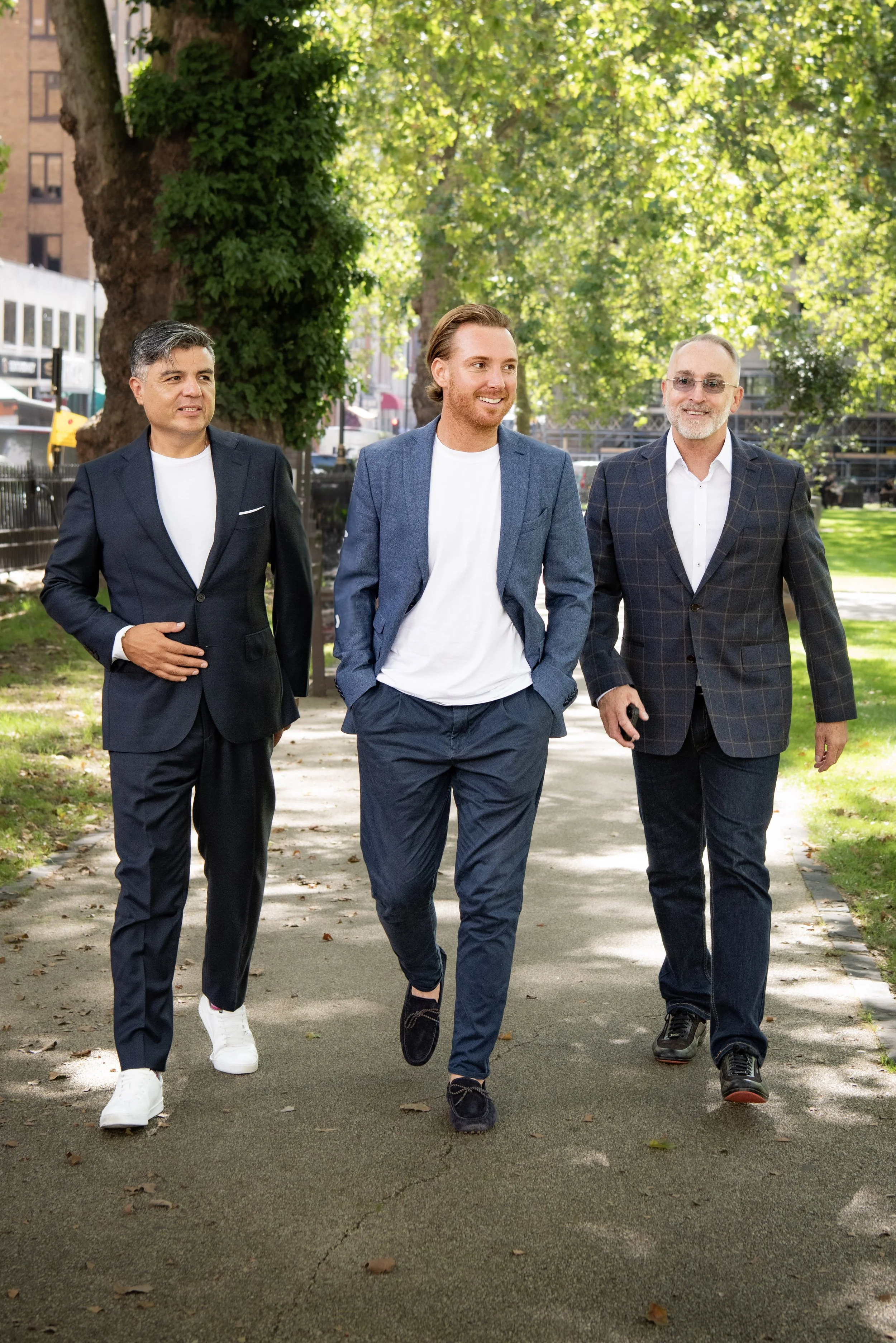 Three men in business attire walking together on a park sidewalk surrounded by green trees and urban buildings.