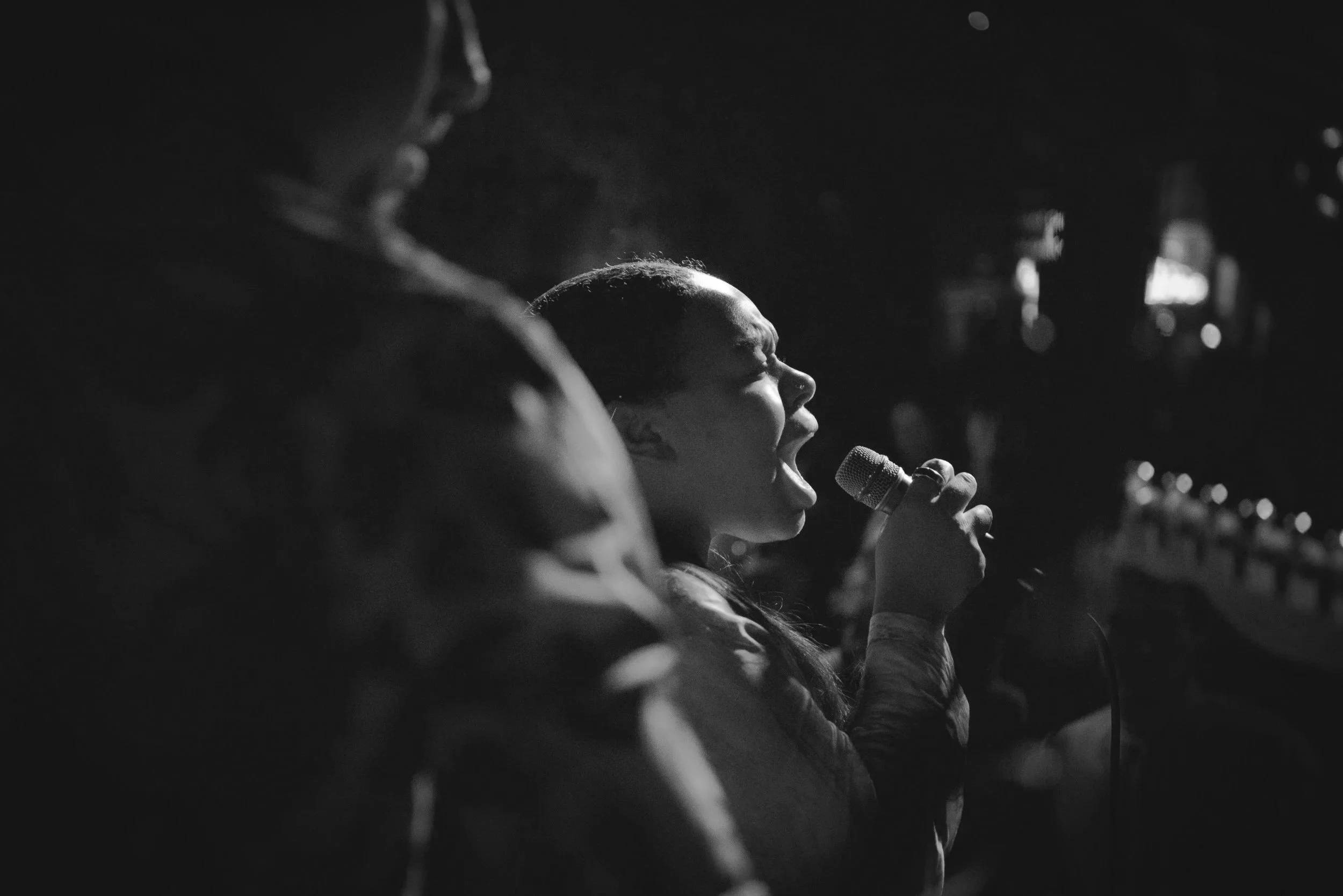 A young woman singing into a microphone, with her eyes closed and a passionate expression, in a dimly lit environment.