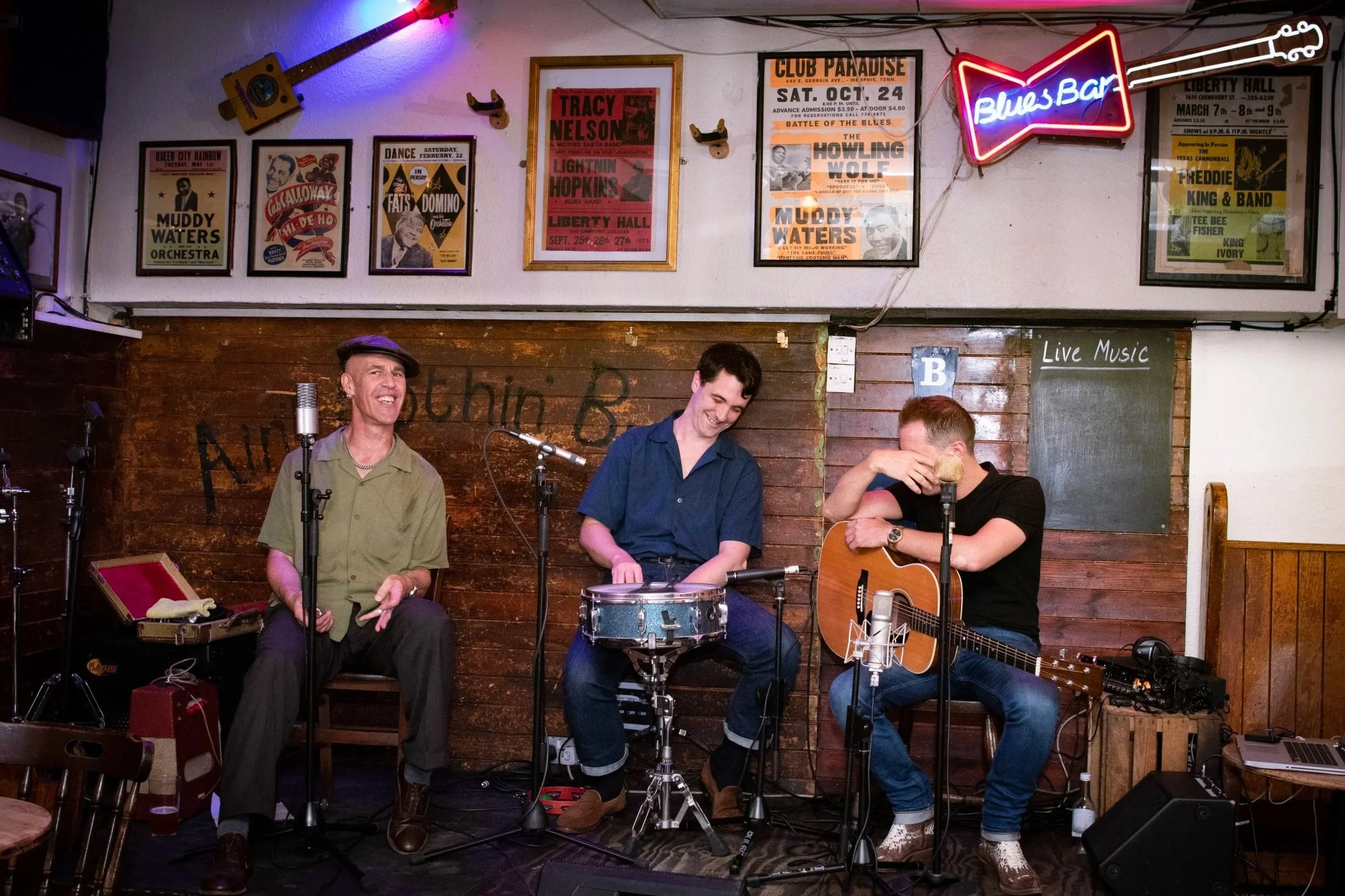 Three male musicians performing live music on stage at a bar or club, with posters on the wall, a neon sign reading 'Blues Bar', and musical equipment around them.
