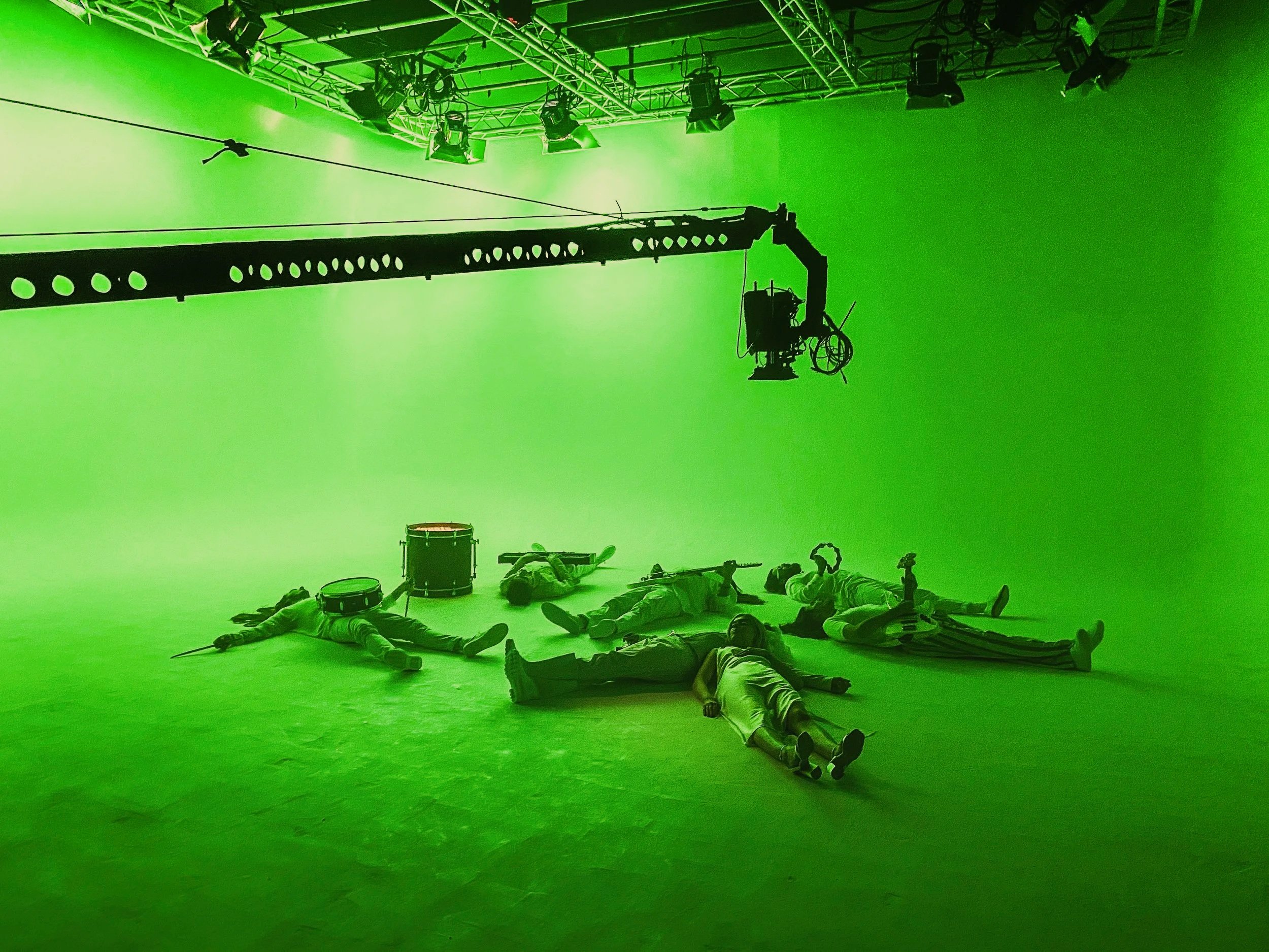 A green screen room with a camera crane overhead and several actors lying on the floor in costumes, with musical instruments around them.