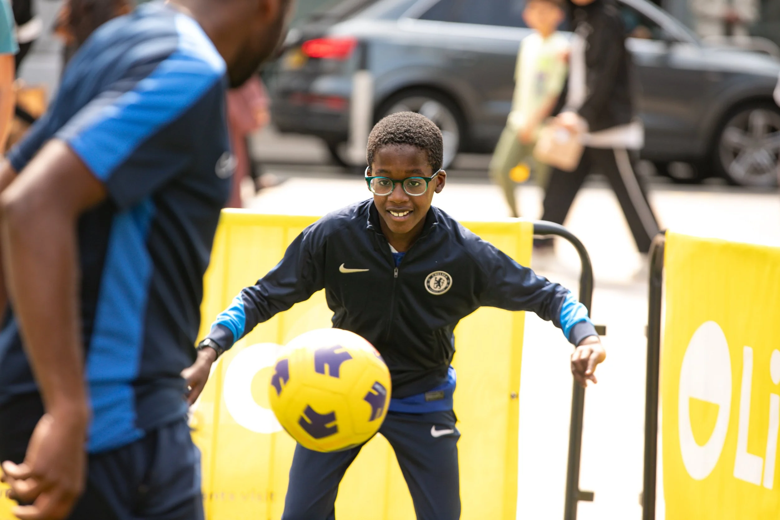 A young boy playing soccer outdoors, wearing glasses and a Chelsea Football Club jacket, with a yellow and purple soccer ball, near yellow barriers, with blurred people and cars in the background.