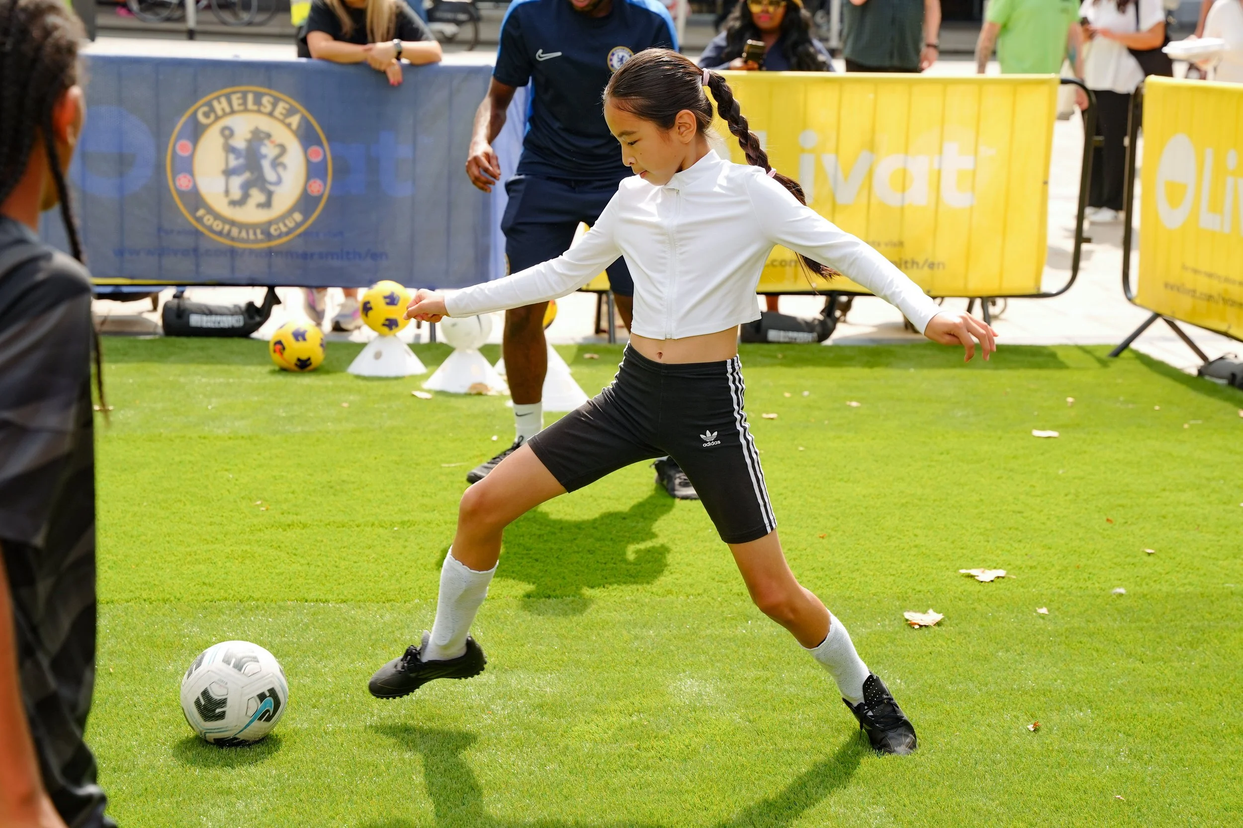 Young girl kicking a soccer ball on a grass field during a public event, with spectators and Chelsea Football Club banners in the background.