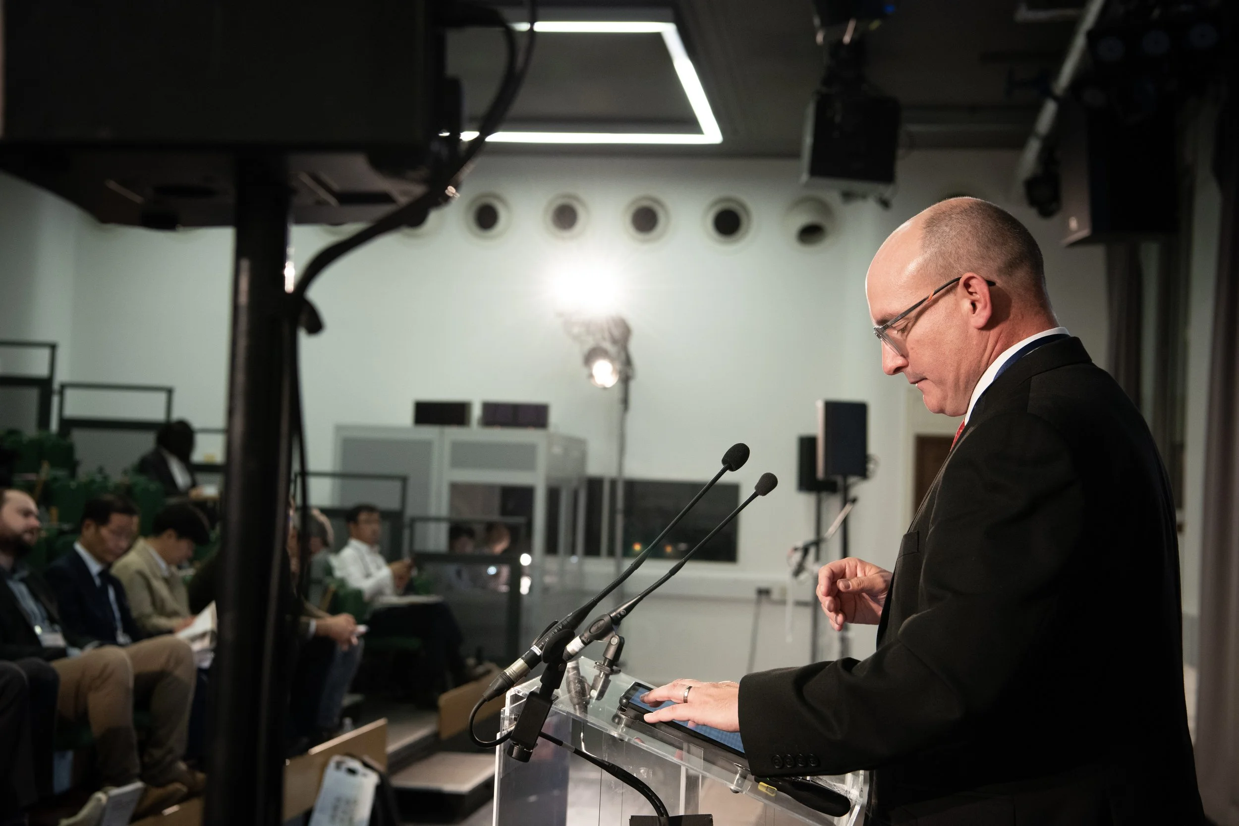 A man in a black suit and glasses giving a speech at a podium with microphones, in front of a seated audience in a conference room.