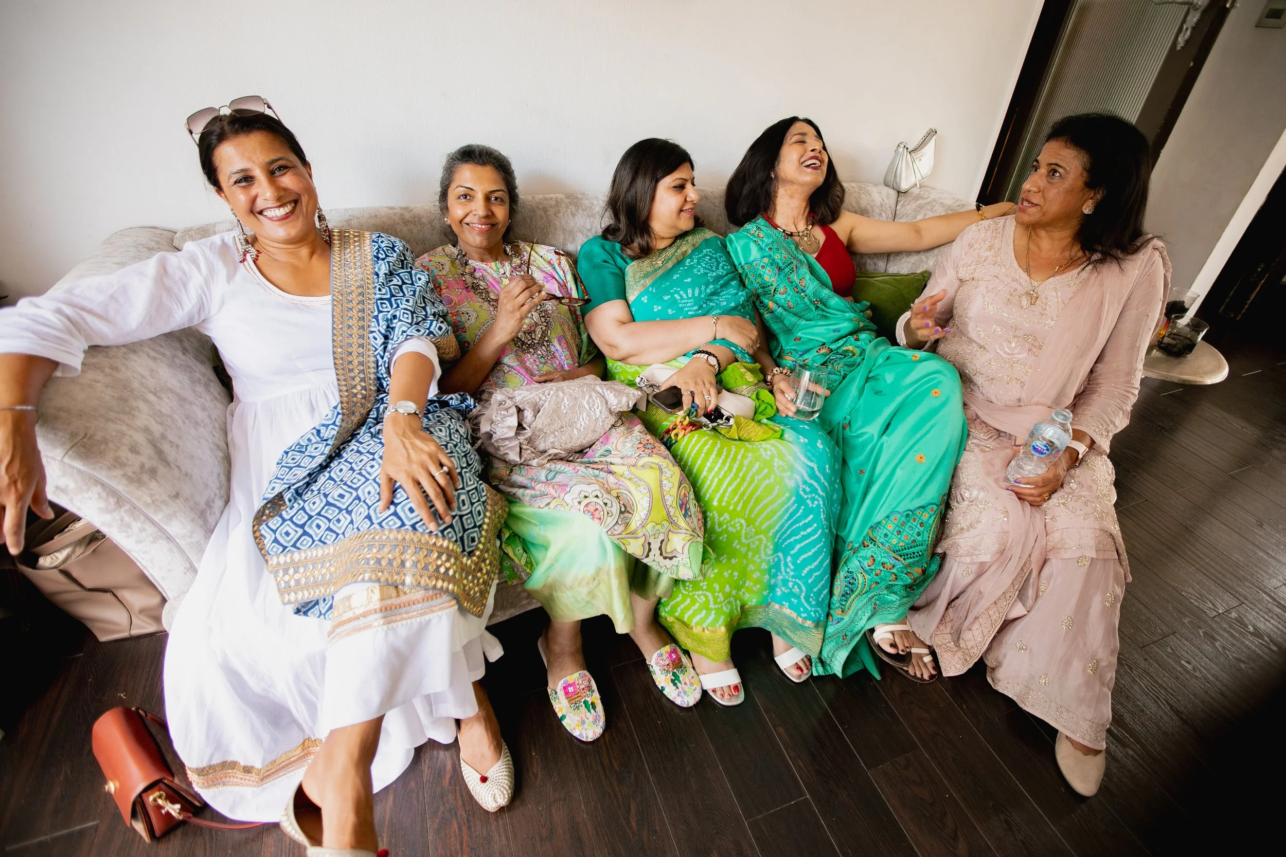 Five women of South Asian descent sitting and smiling on a beige couch, wearing colorful traditional dresses, in a living room.