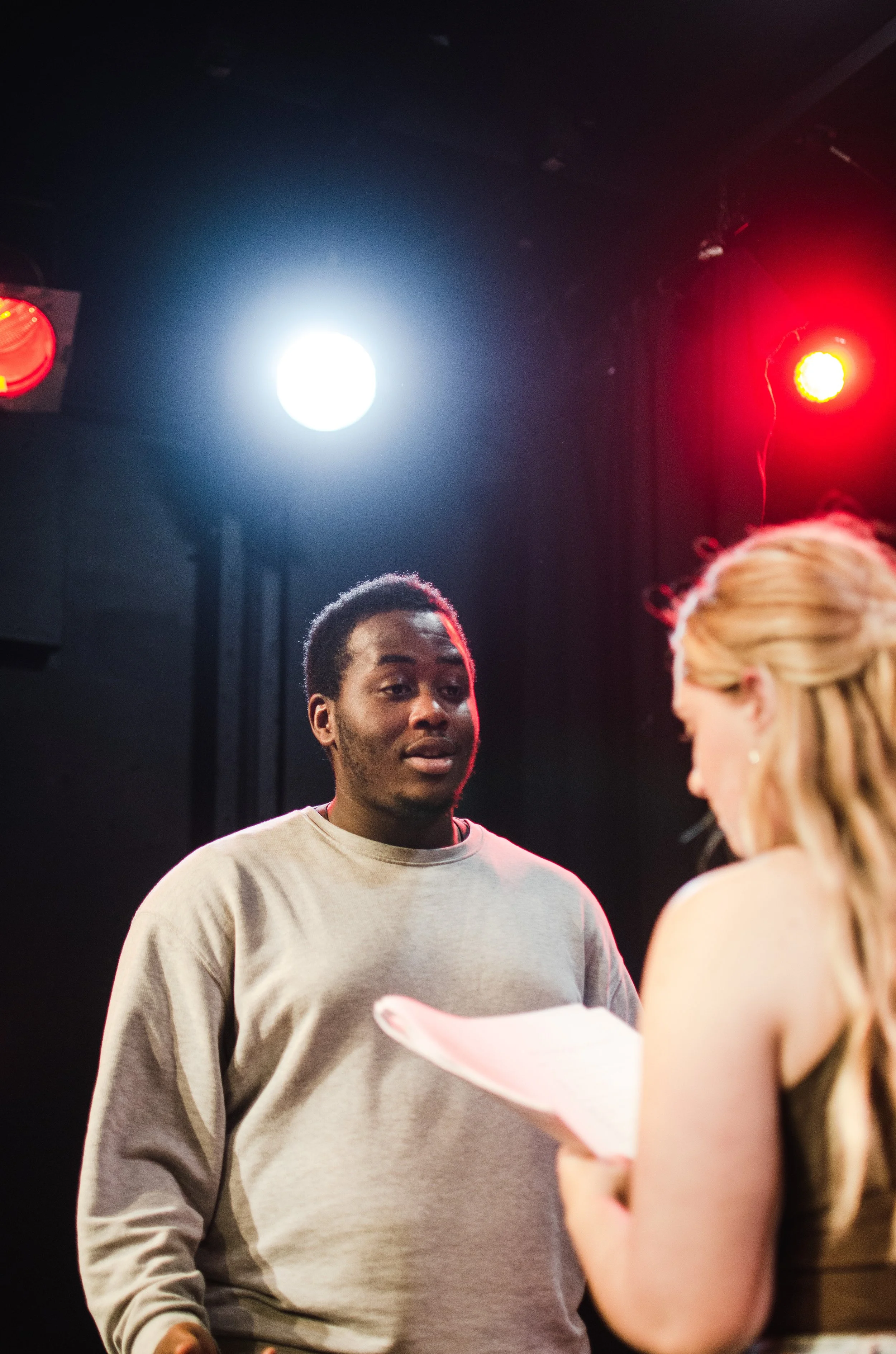 A scene from a theater rehearsal or acting class showing a man talking to a woman, with stage lights overhead.