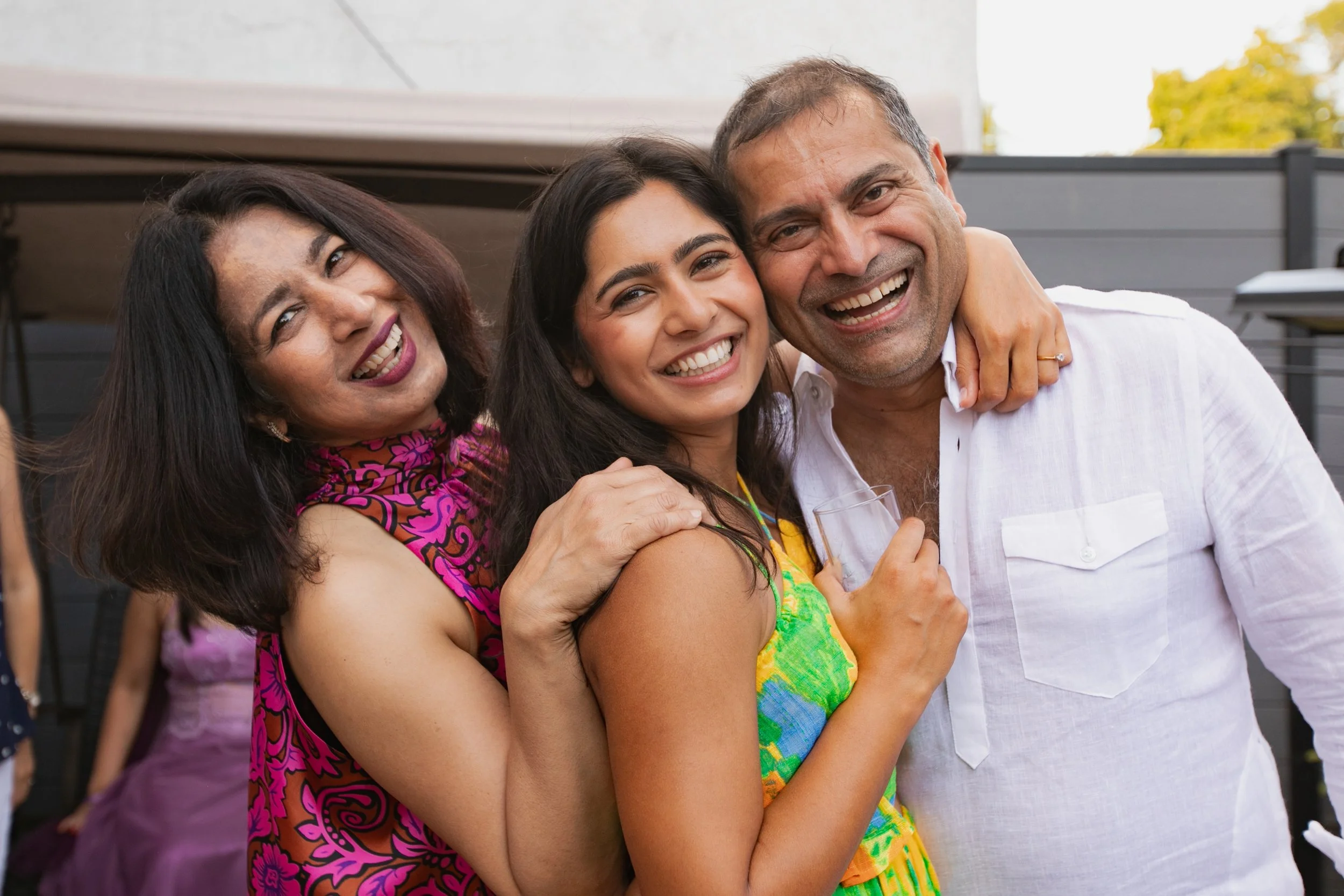 Three people smiling, celebrating, and hugging outdoors at a social gathering.