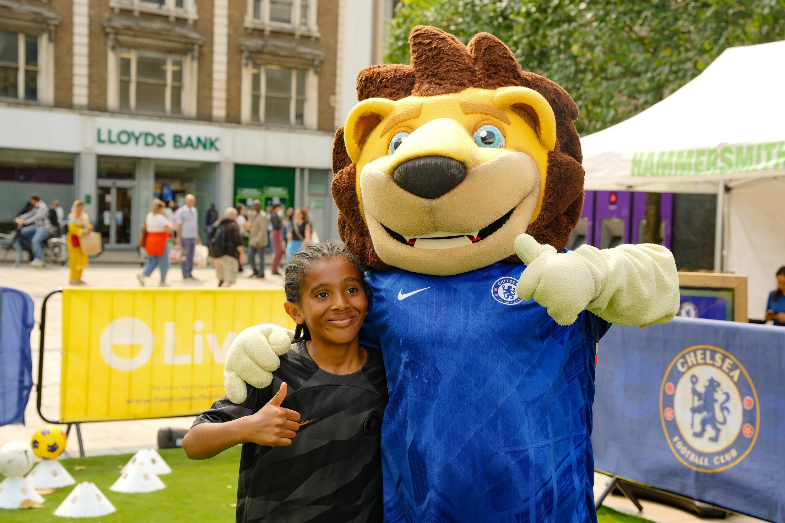 A young girl with braided hair posing with a lion mascot wearing a Chelsea Football Club jersey at an outdoor event, with a street scene and people in the background.