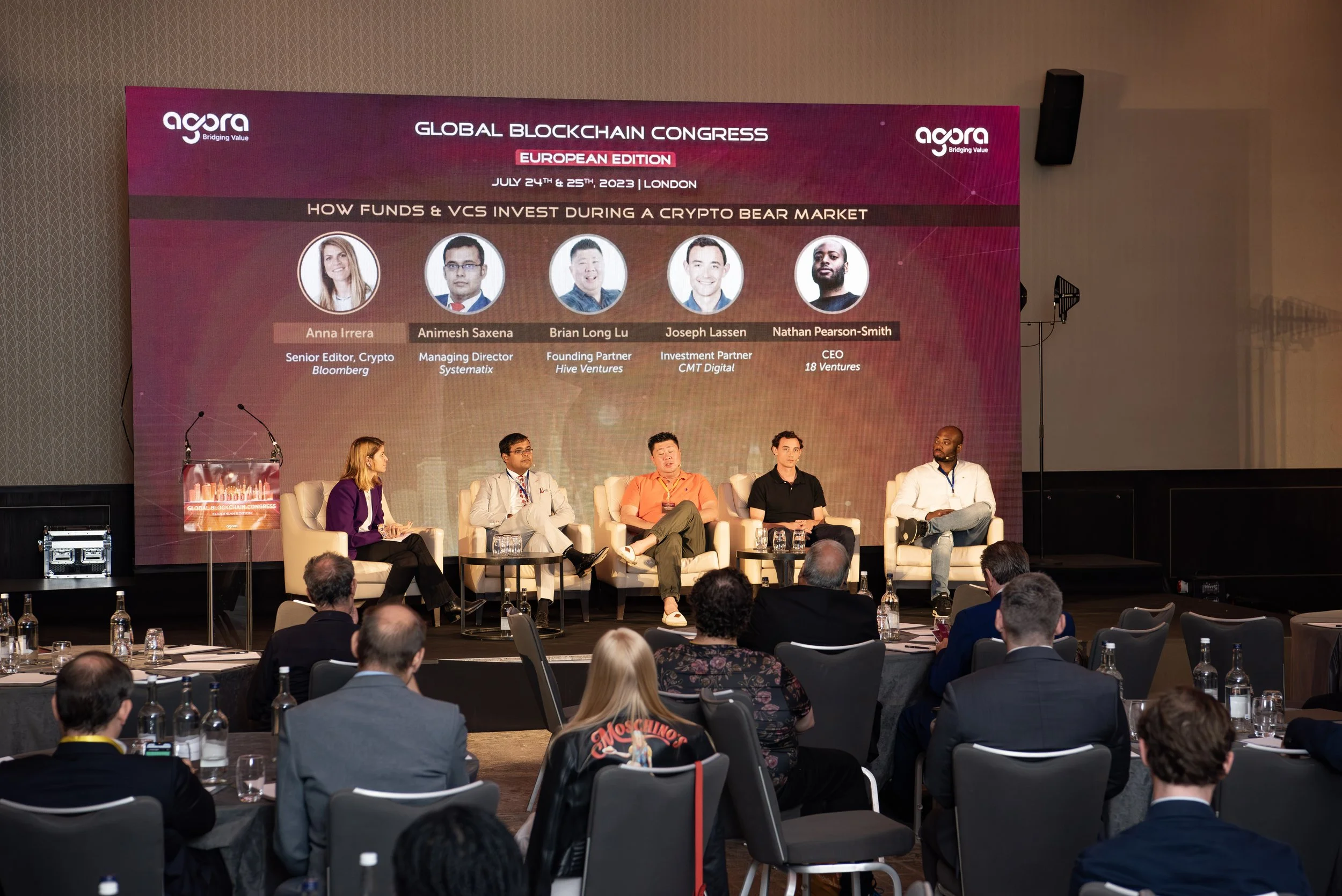 A panel discussion at the Global Blockchain Congress, European Edition, held in London on July 24-25, 2023. The stage has five speakers seated in armchairs, with a large screen behind them displaying their names, titles, and photos. The audience is s