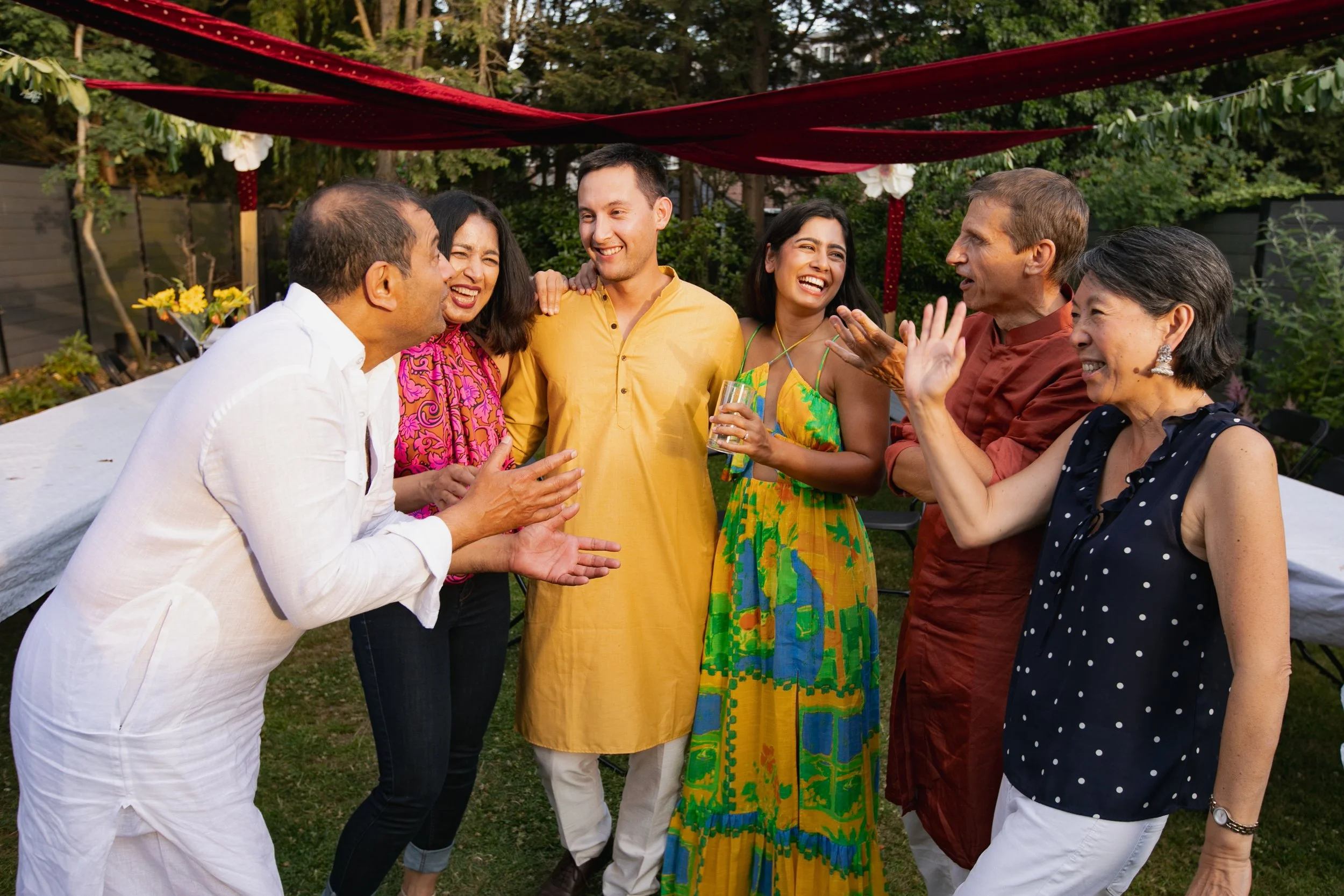 Group of people laughing and talking at a garden party with decorated outdoor space and trees in the background.