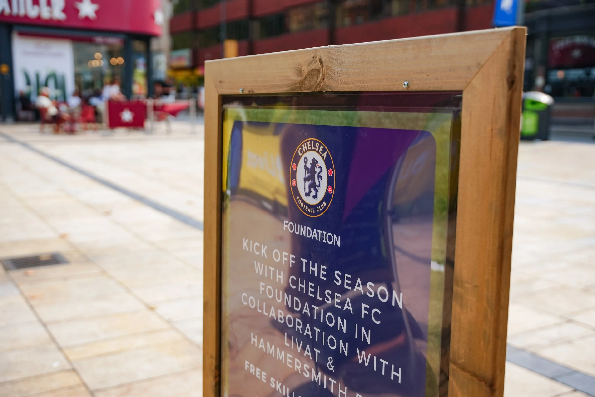 A wooden-framed sign on a busy city sidewalk with the Chelsea Football Club logo, advertising a foundation event for Chelsea FC.
