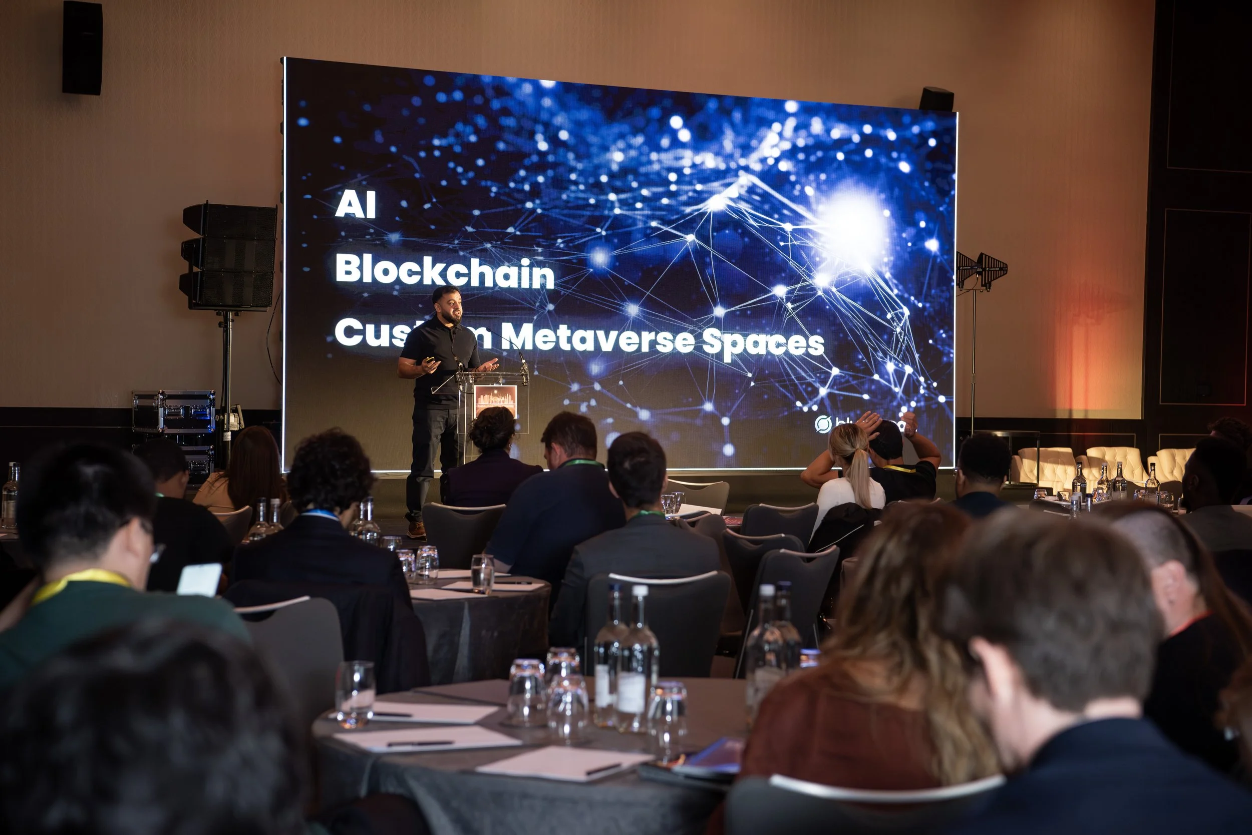 A speaker presenting on stage at a conference with a large screen that reads 'AI Blockchain Custom Metaverse Spaces.' The audience is seated at round tables, listening attentively.