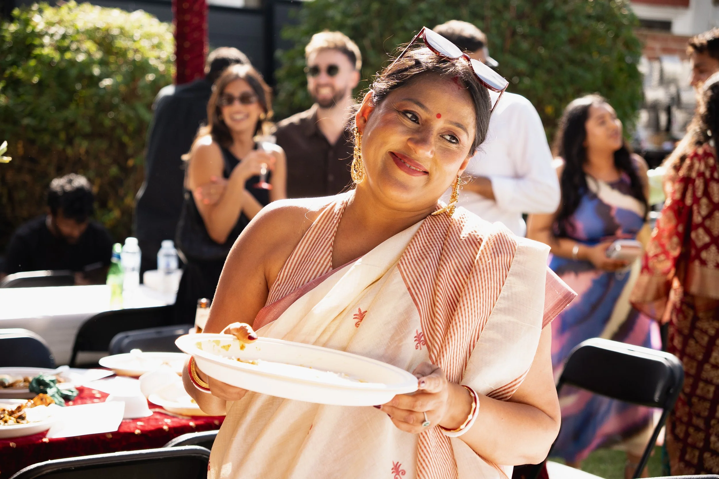 A woman in traditional Indian attire holding a white plate at an outdoor gathering, smiling. Other people are in the background, some holding drinks, in a garden setting with trees and tables.