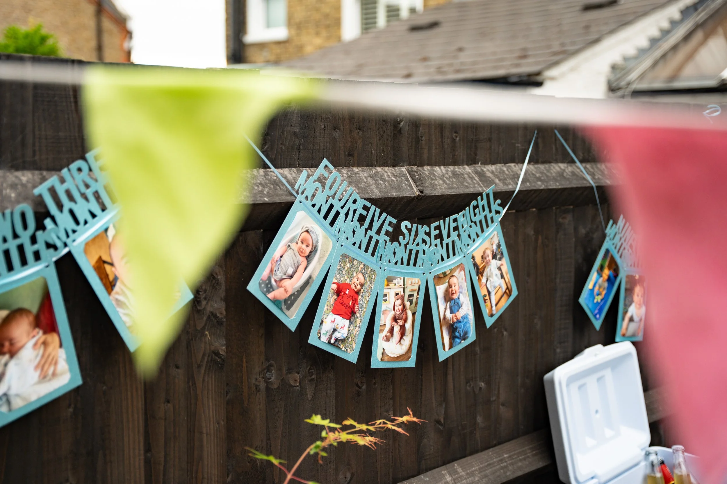 Photo of a string banner featuring multiple printed photographs of a young girl, attached with blue paper cutouts that read 'THREE YEARS OLD' and other words. The banner is hung on a wooden fence, with a house and some greenery visible in the backgro