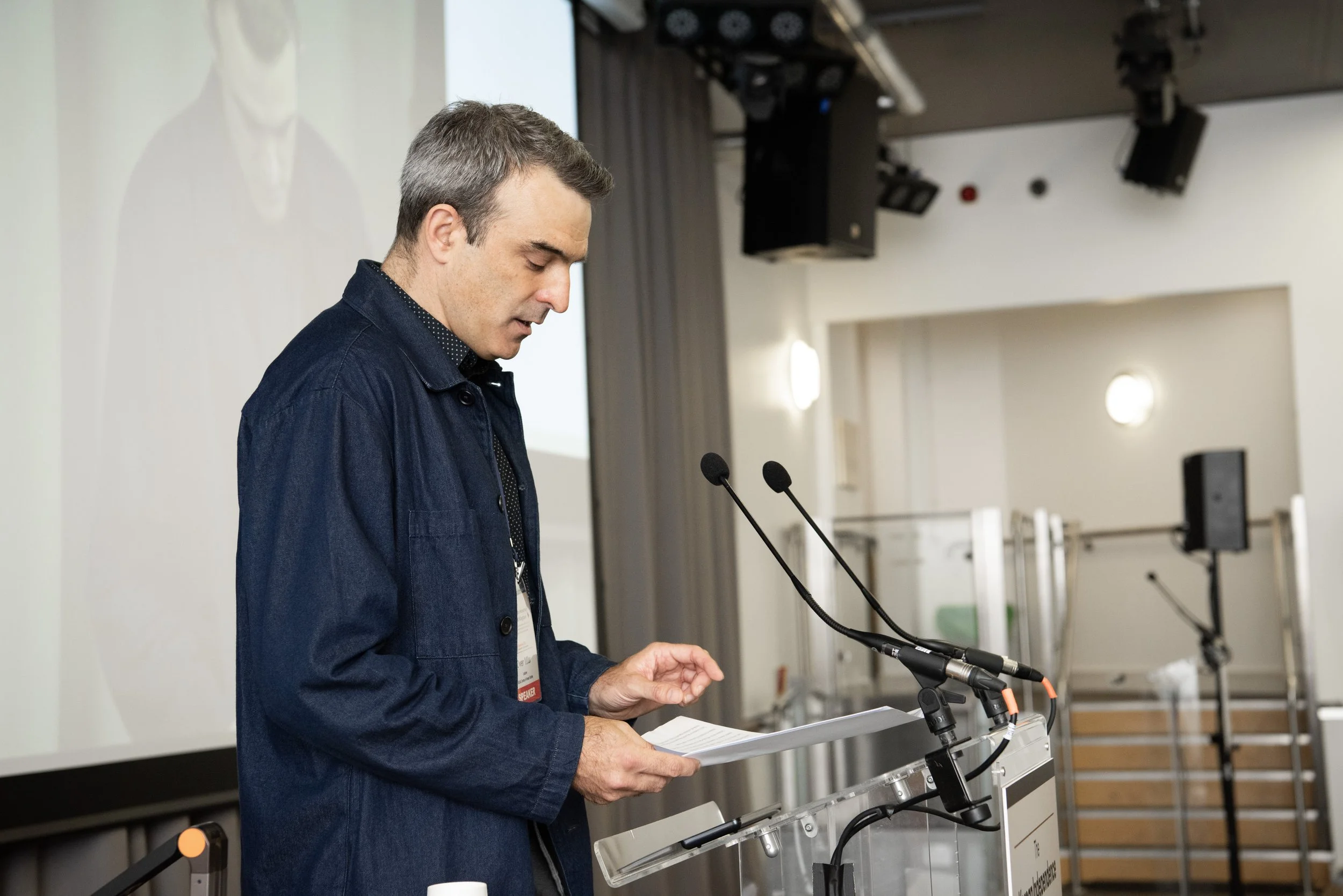 A man standing at a podium with two microphones, reading from a paper in a conference room with a large screen behind him.