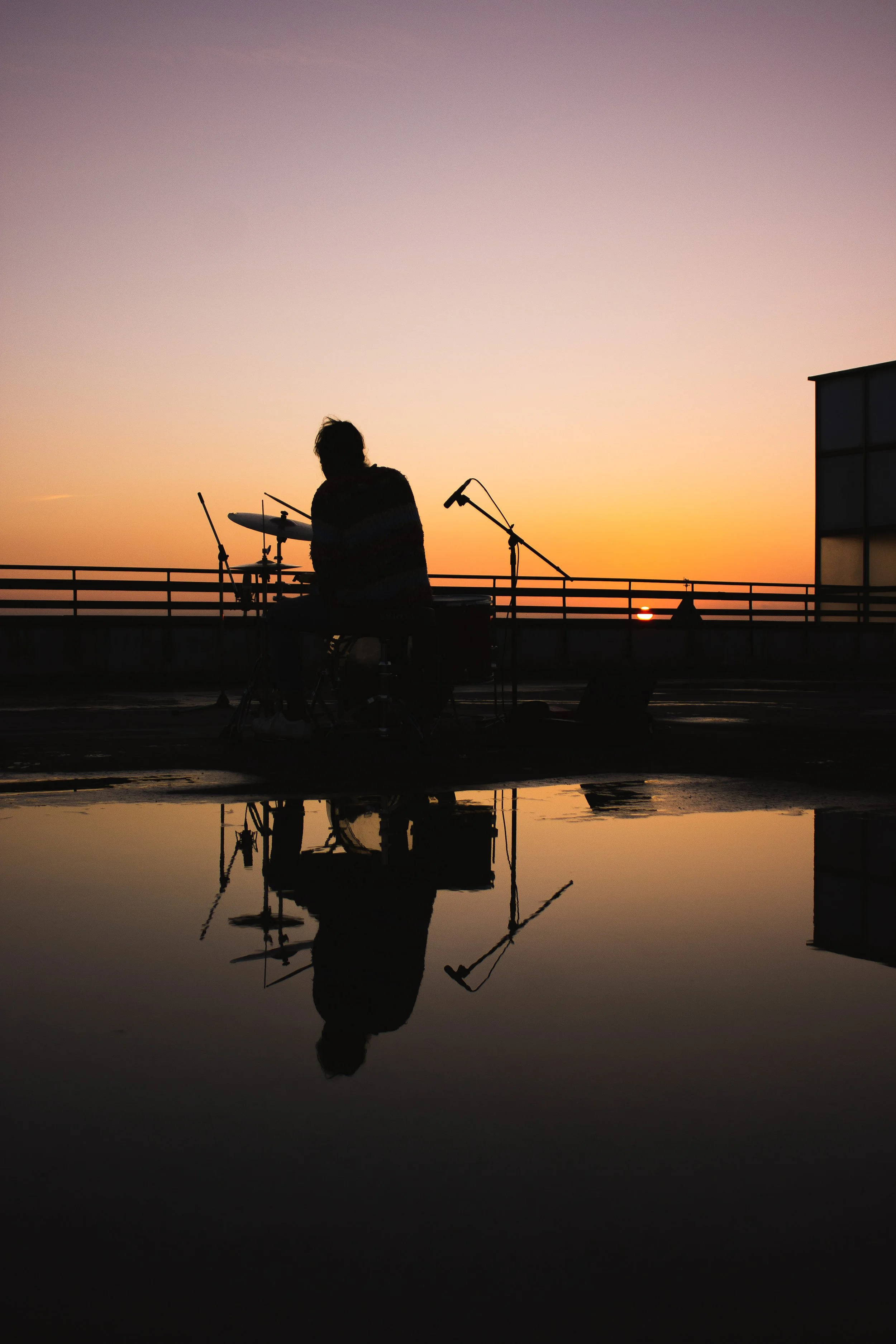 Silhouette of a person sitting with a drum set and microphone on a rooftop at sunset, with their reflection visible in a puddle on the ground.