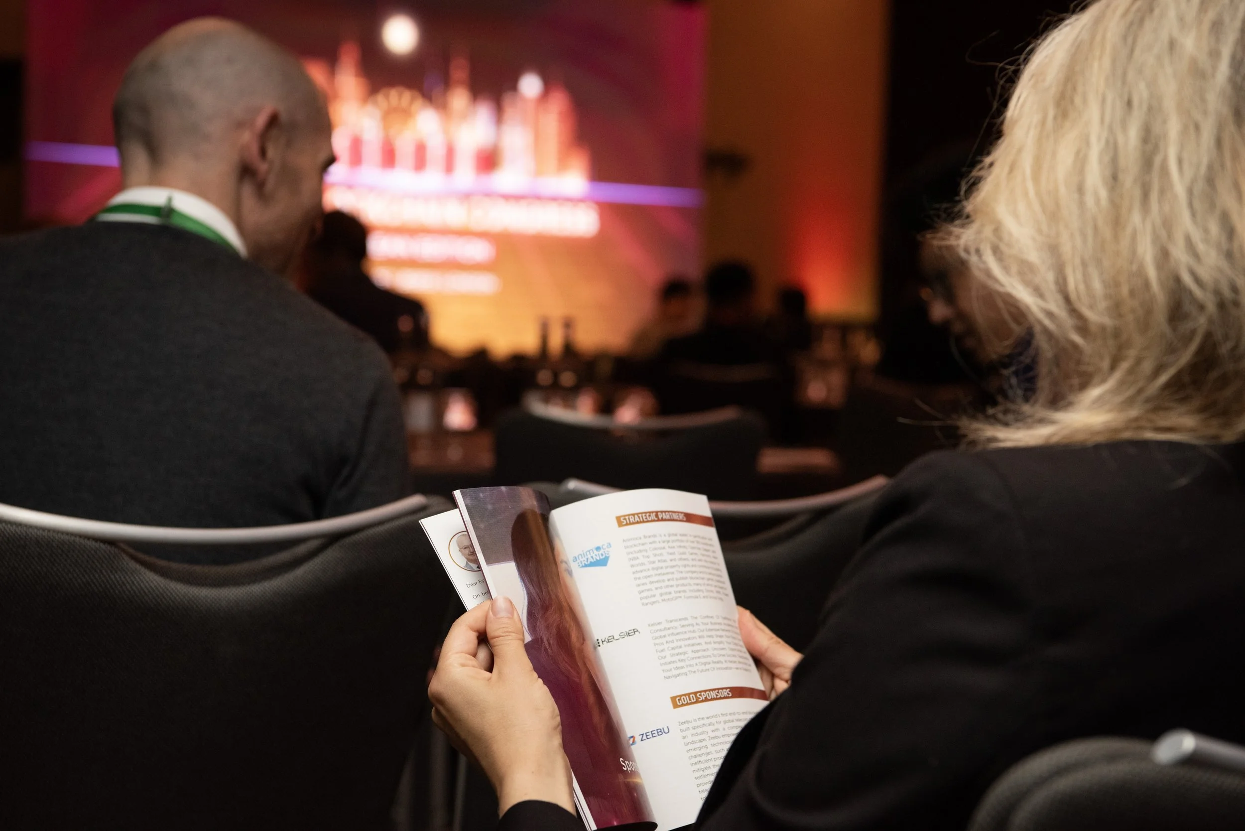 A woman reading a program booklet at a conference or event, with a stage and audience visible in the background.