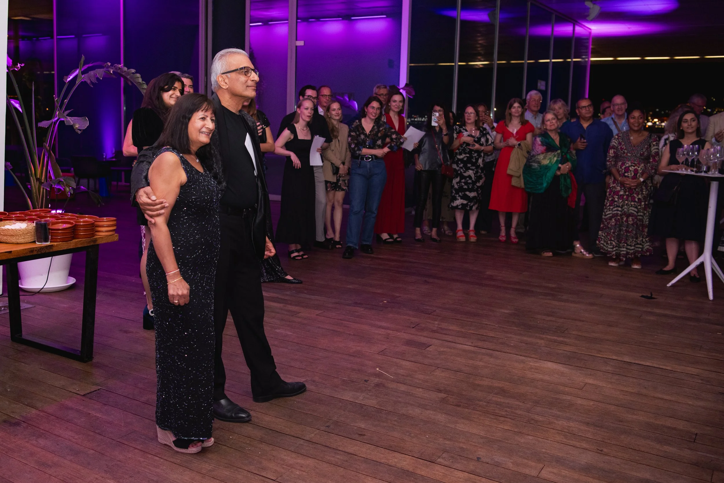 A group of people at an indoor event, with two individuals in the foreground, one woman in a sparkly black dress and a man in black attire, standing closely together while others stand in the background, some holding papers, in a room with glass wall