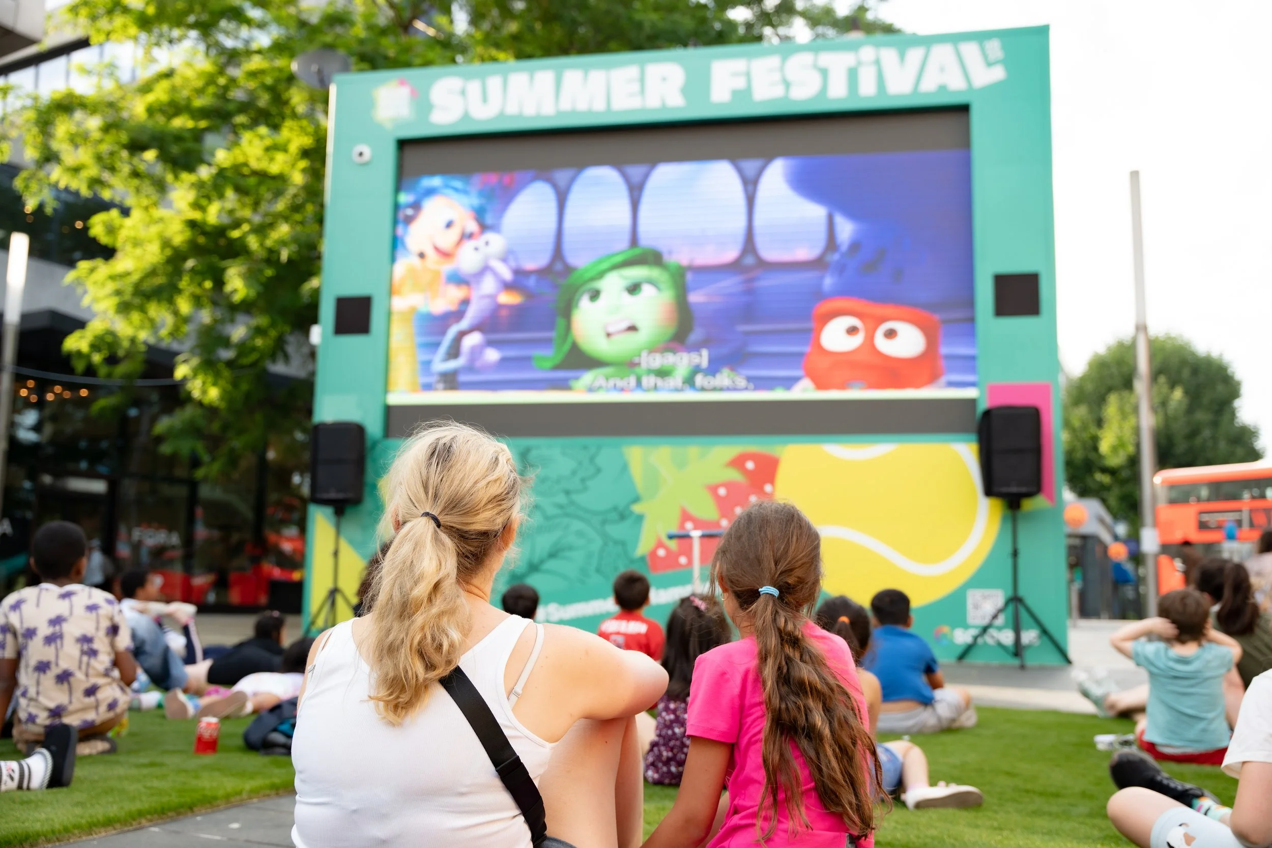 People watching an outdoor movie screening on a large screen in a park, with children and adults sitting on the grass during a summer festival.