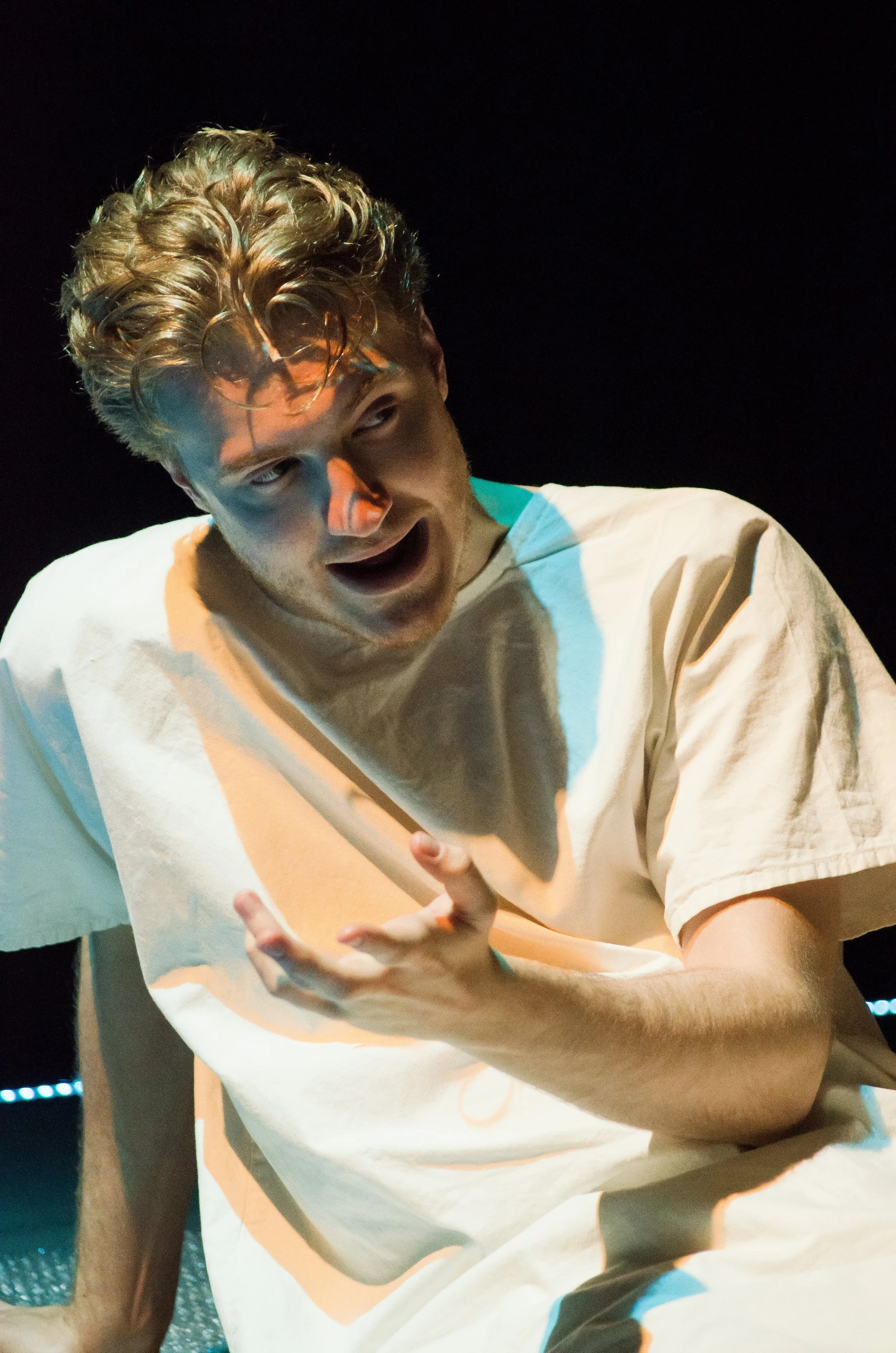 A young man with curly blonde hair acting dramatically on stage, wearing a white t-shirt, with a dark background.