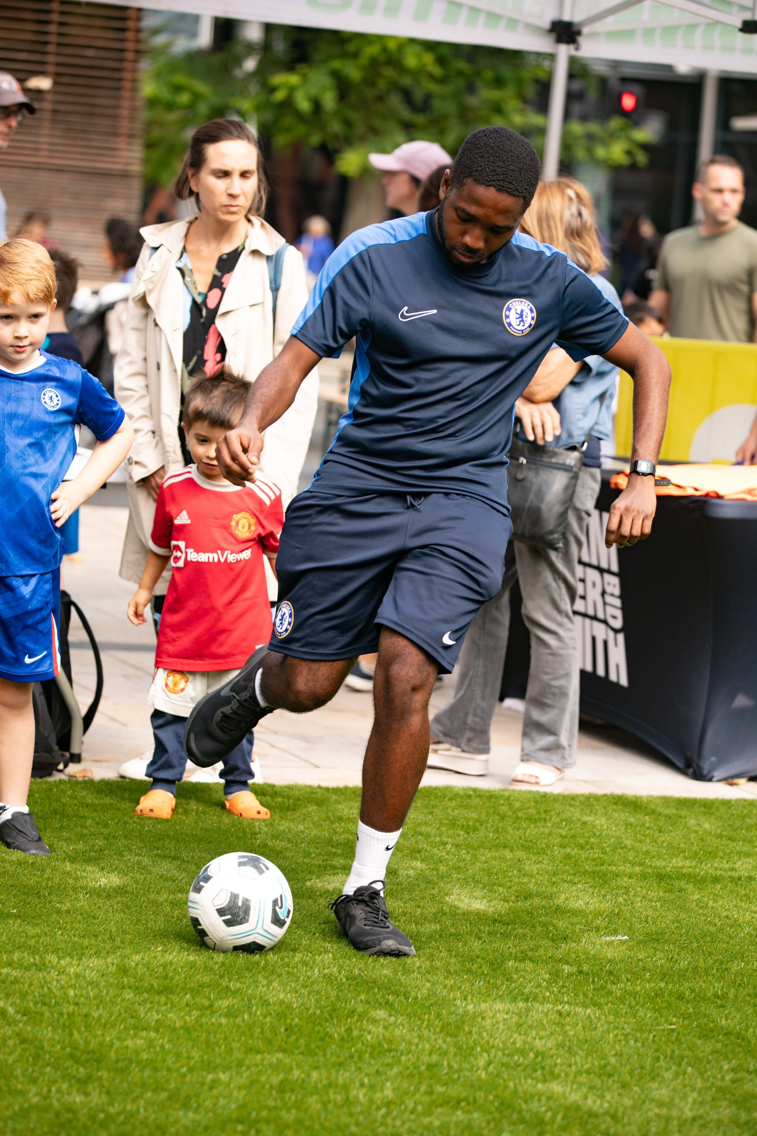 A man wearing a Chelsea football jersey playing soccer on a small field, with children and adults watching.
