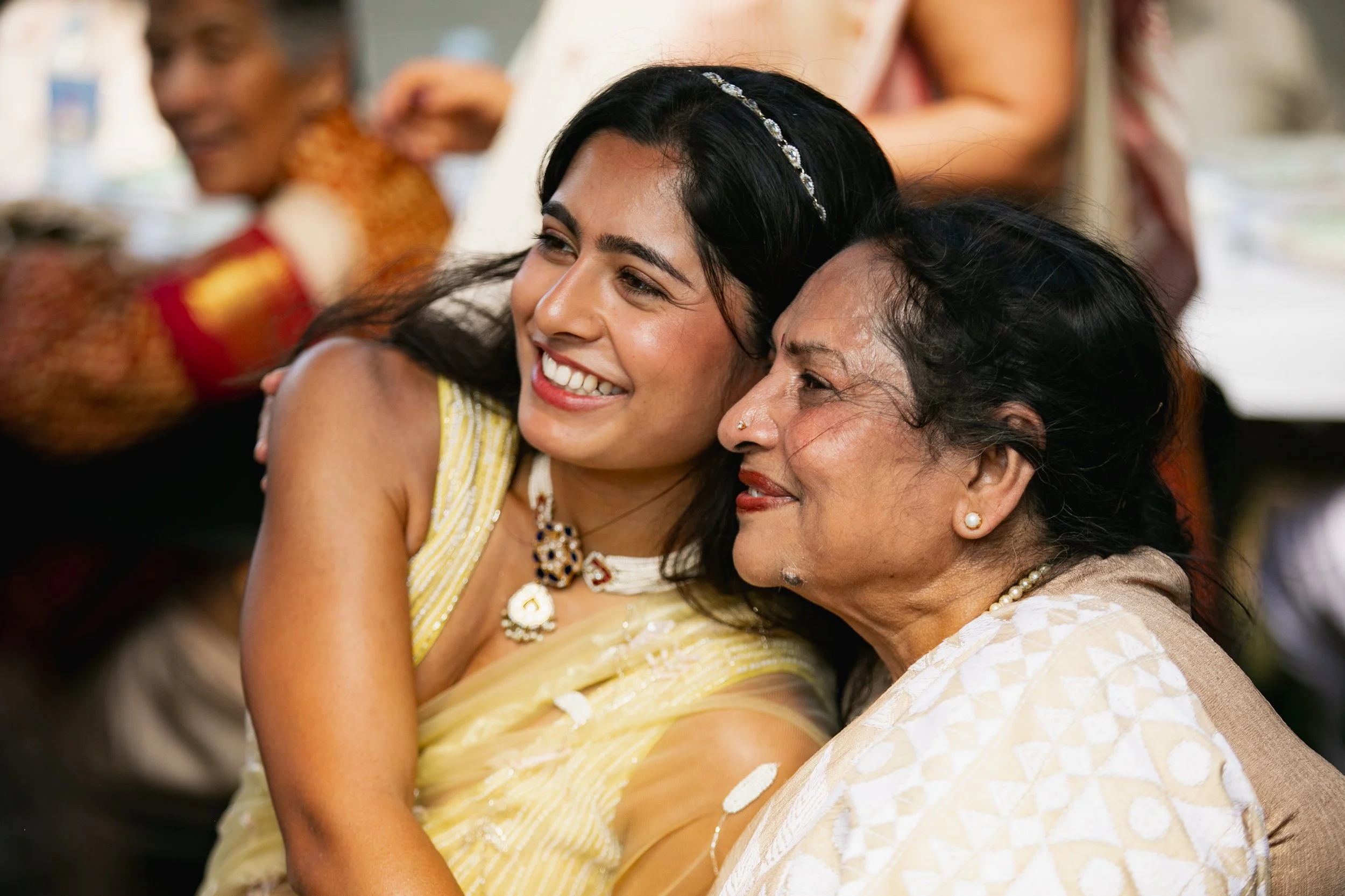 Two women embracing and smiling at a celebration or gathering, with other people visible in the background.