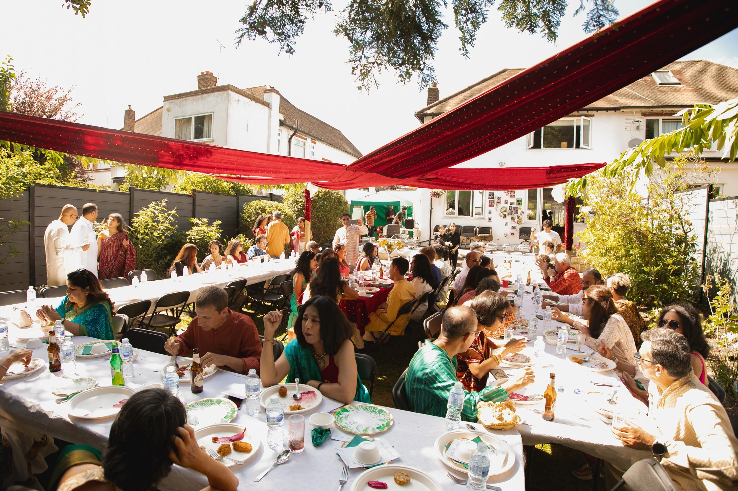 An outdoor gathering with many people sitting at long dining tables under red fabric drapes, in a backyard with houses and trees in the background.