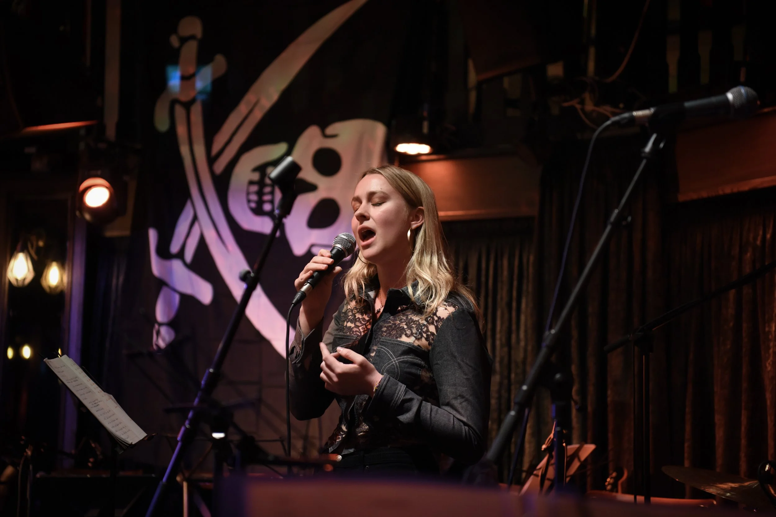 A woman singing into a microphone during a live performance on stage with a dark background and a skull musical note logo.