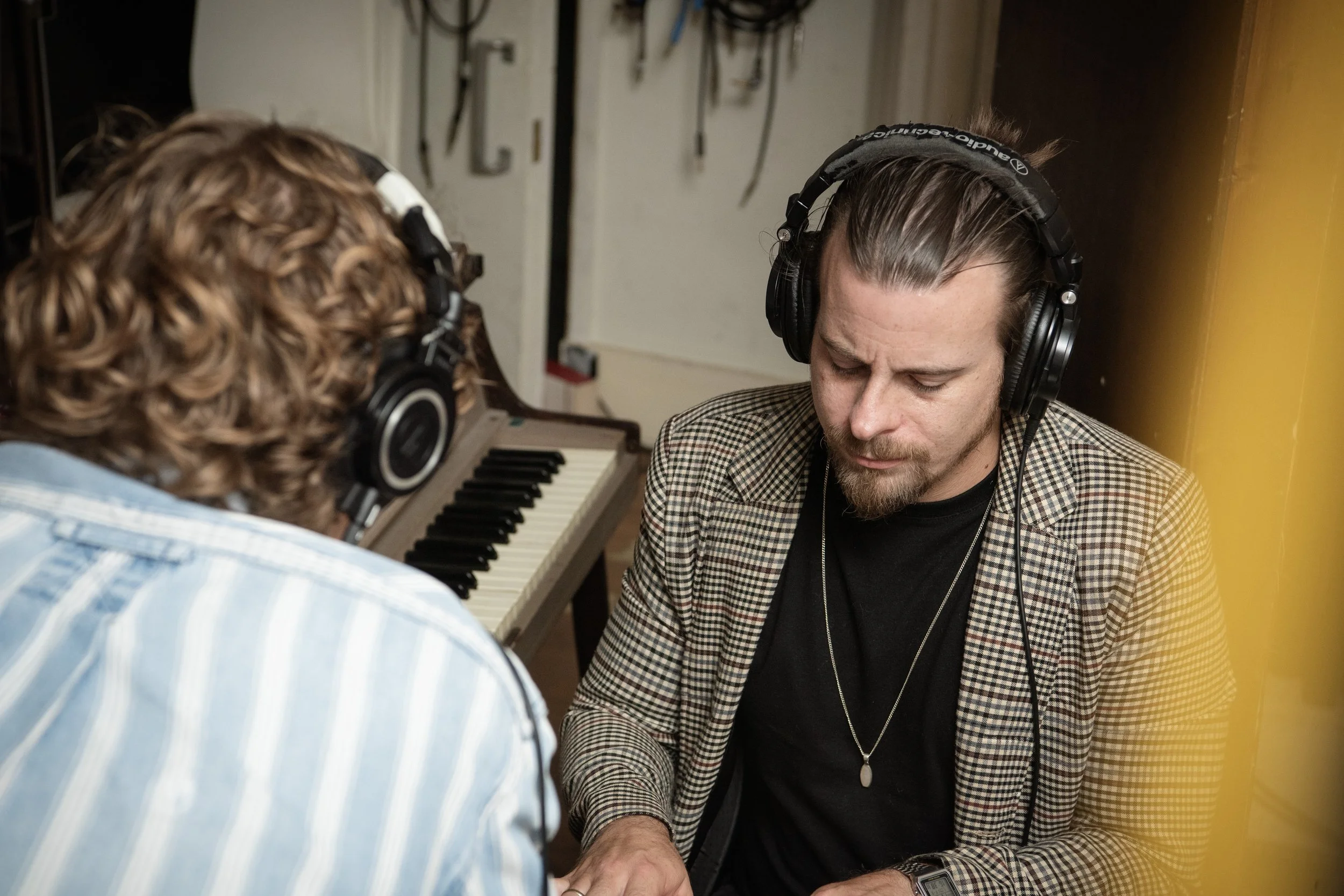 Two men wearing headphones, one with curly hair and the other with straight hair, sitting in front of a keyboard in a music studio.