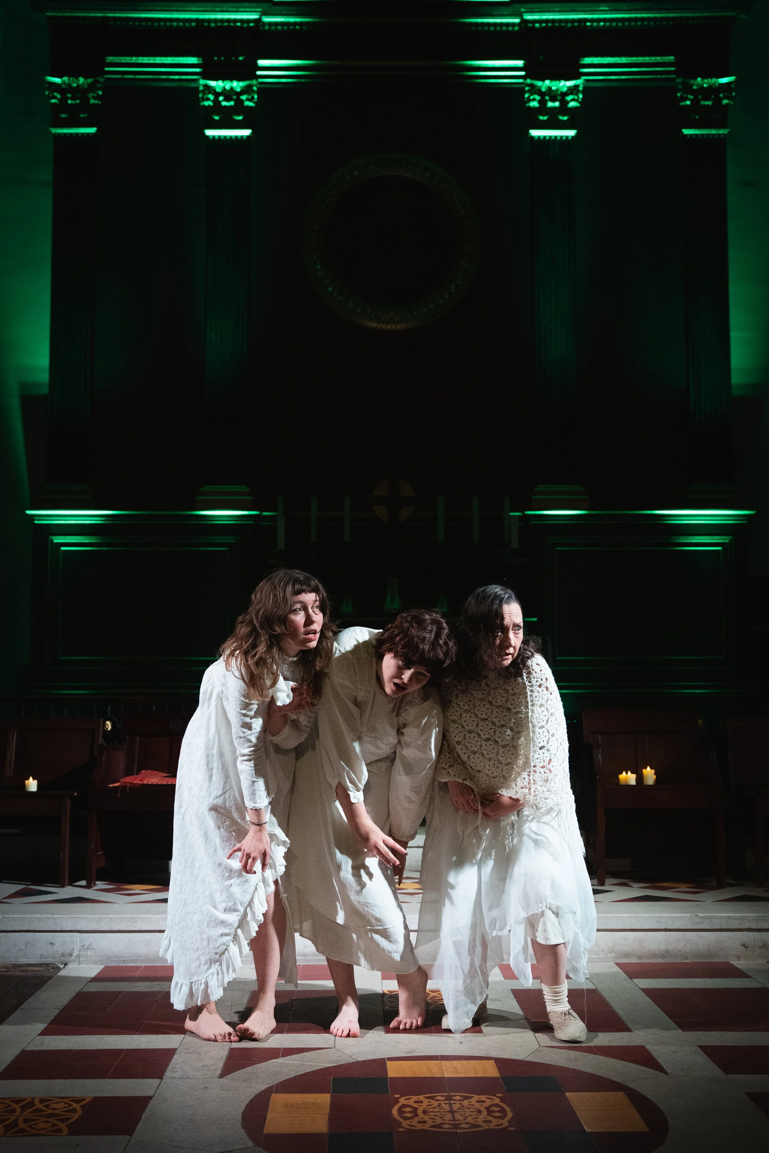 Four women dressed in white garments in a theatrical or staged scene, standing in a dimly lit room with candles and a dark backdrop.