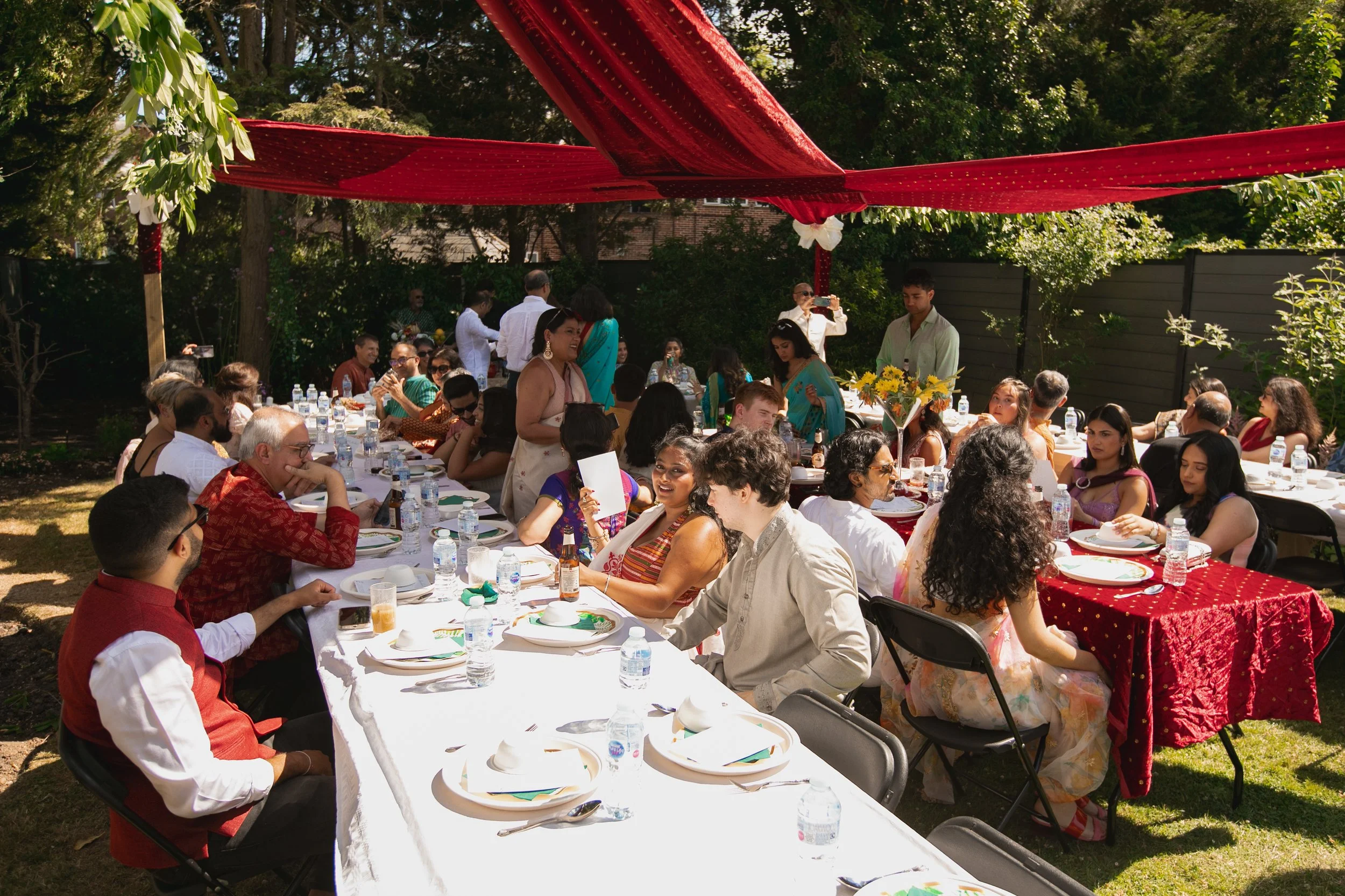 People gathered around dining tables outdoors for a celebration under a red tent, with trees and greenery in the background.