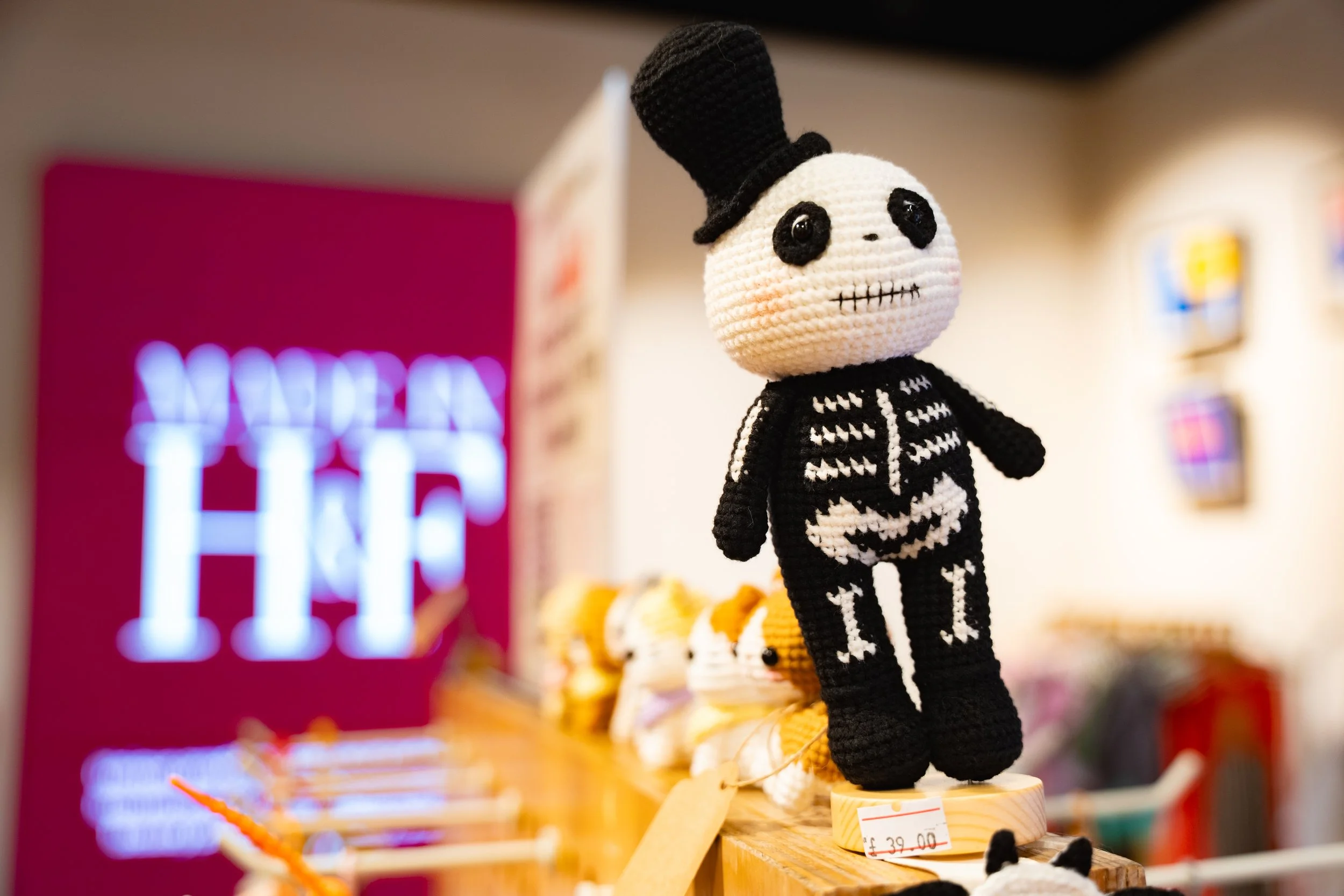 A crocheted skeleton doll with a skull face, black eyes, and a stitched mouth, dressed in a skeleton costume and black top hat, standing on a wooden display shelf in a store.